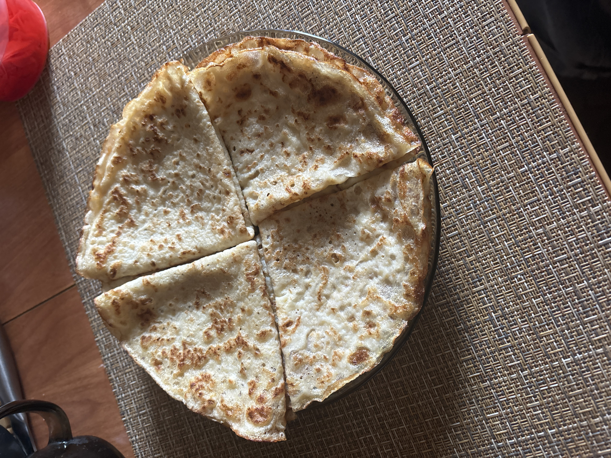 A top-down shot shows a clear glass plate filled with four sections of sliced pancakes, arranged on a woven beige placemat. The placemat sits on a wooden surface, with glimpses of other objects: a red container and a black kettle. The pancakes have a light brown, speckled appearance, and are cut into triangular portions. The placemat features a regular, interwoven pattern. The image was taken in Chaikovsky, Russia.