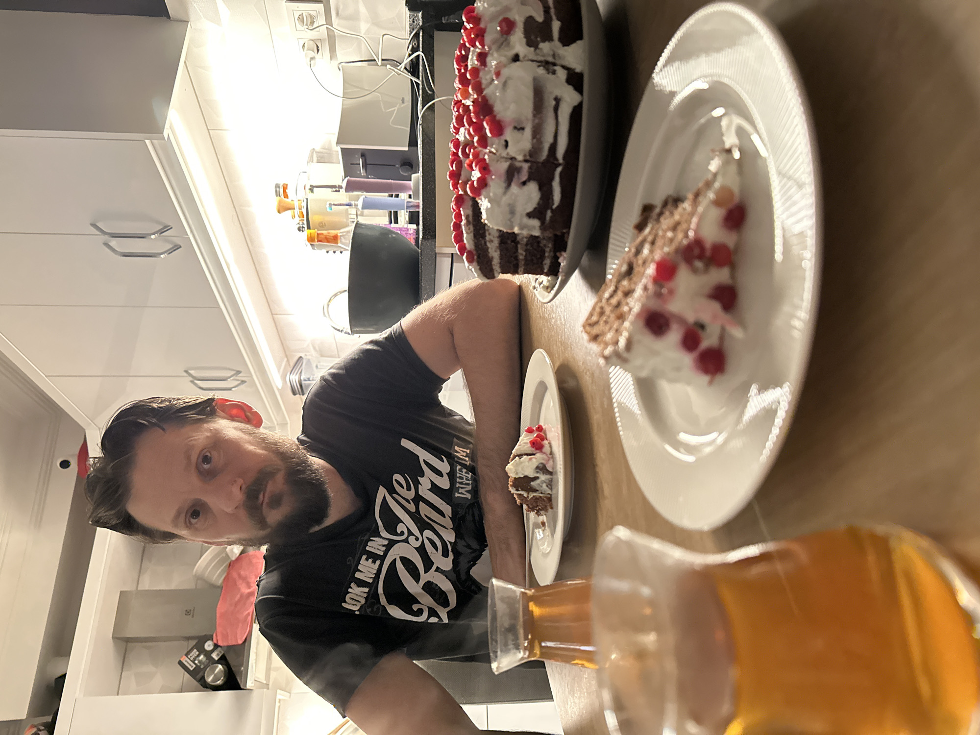 A man with a beard is in a kitchen, posing with a chocolate cake decorated with white frosting and red berries. A slice of cake is on a smaller plate near him, and a larger slice is on a plate next to the whole cake. A glass of amber-colored liquid is in the foreground. The kitchen has white cabinets and countertops. The location is not discernible as Fethiye, Turkey from this image alone.