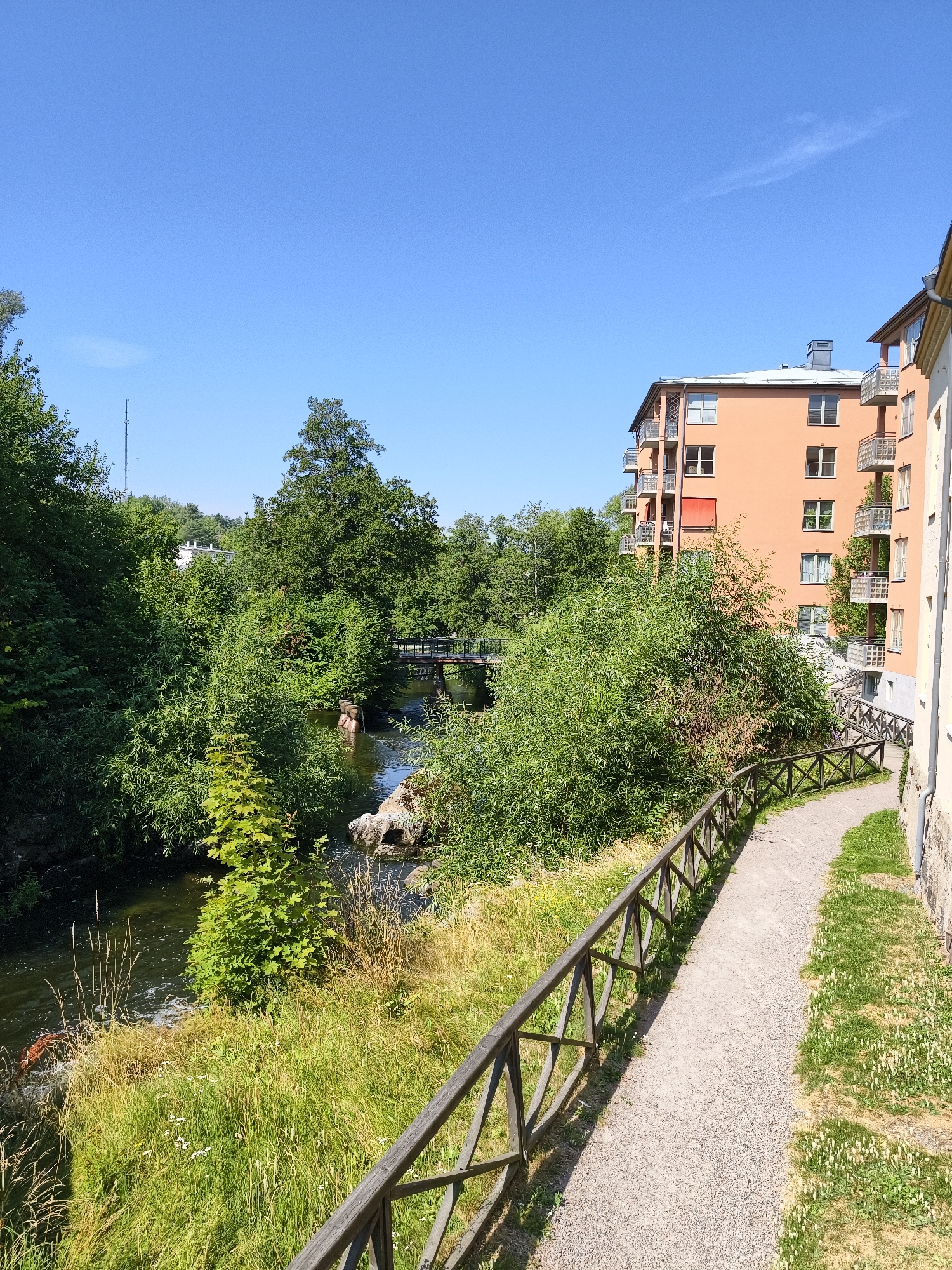 The image shows a scenic view of the Motala ström river in Linköping, Sweden, on a clear, sunny day. A paved walking path lined with a rustic wooden fence meanders alongside the riverbank. Lush green trees and vegetation flank the water, creating a serene natural setting. In the background, an orange apartment building with multiple balconies stands out against the blue sky, suggesting an urban environment blending with nature. A pedestrian bridge can be seen crossing the river further upstream, and a tall radio tower is visible in the distance, peeking above the trees. The scene is peaceful and depicts a popular spot for recreation and enjoying the outdoors in the city.