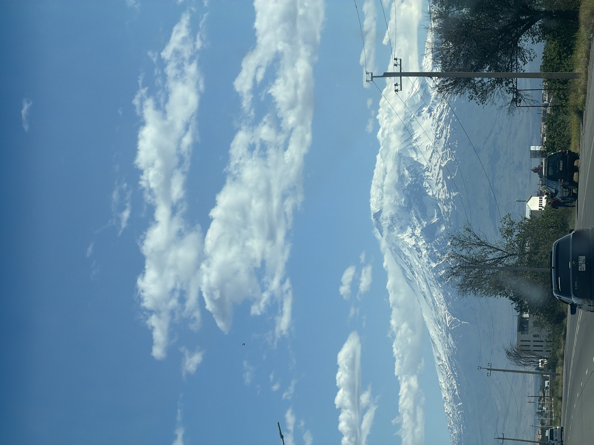 The image shows a view of a large, snow-capped mountain range under a blue sky with scattered clouds. The view is from a roadside, with utility poles visible along the side of the road. Cars are parked along the roadside. The location appears to be within the city of Unknown, Unknown. A few buildings are seen in the distance. The camera is oriented vertically.