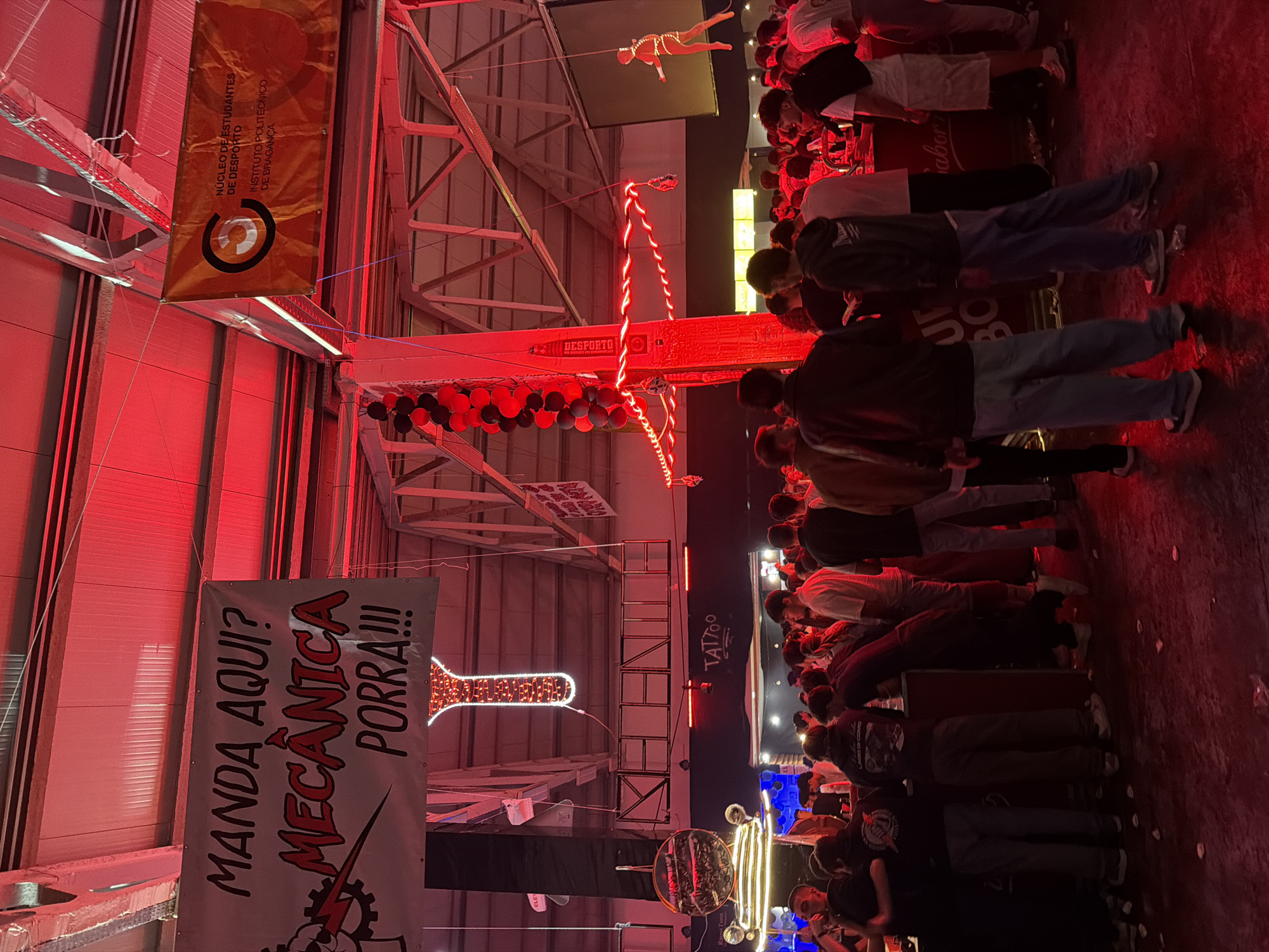 The image shows a crowded indoor event, possibly a student or university-related gathering, likely taking place in the city of Bragança, Portugal. The scene is bathed in red lighting. Numerous individuals are present, mostly young people. Banners are visible, one advertising a "MECÂNICA PORRA!!!" event with related imagery. Another banner identifies the "NÚCLEO DE ESTUDANTES DE DESPORTO" (Sports Student Association) of the "INSTITUTO POLITÉCNICO DE BRAGANÇA" (Polytechnic Institute of Bragança). There is a lit-up decorative wrench. Some people are walking around. Above the crowd, a figure hangs from a cable.