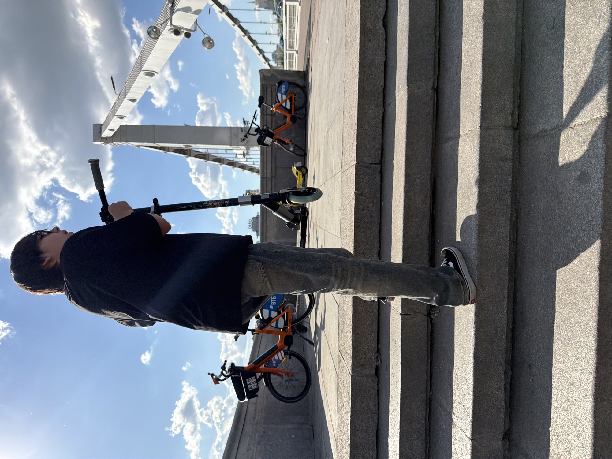 An outdoor scene captures a person standing on a paved walkway adjacent to a flight of concrete steps, under a bright, partly cloudy sky. The image is oriented sideways, but if rotated, the person stands on the left, looking towards the left. The individual, dressed in a black t-shirt, jeans, and dark sneakers with white soles, holds an electric scooter with a black frame and a yellow component near its base.
In the background, a large metal bridge structure with visible support beams and overhead lights spans the scene. Parked near the steps are two orange share bikes, one closer to the person and another further in the background. Distinct, long shadows stretch across the steps and the ground, suggesting a low sun angle, indicative of either early morning or late afternoon.
Visible text on one of the orange share bikes includes "ВТБ" (VTB, a Russian bank) and "AA6 281" on a tag attached to its front basket. The electric scooter also displays "OKKO" on its frame. The sky is blue with scattered white cumulus clouds. The setting is Moscow, Russia.