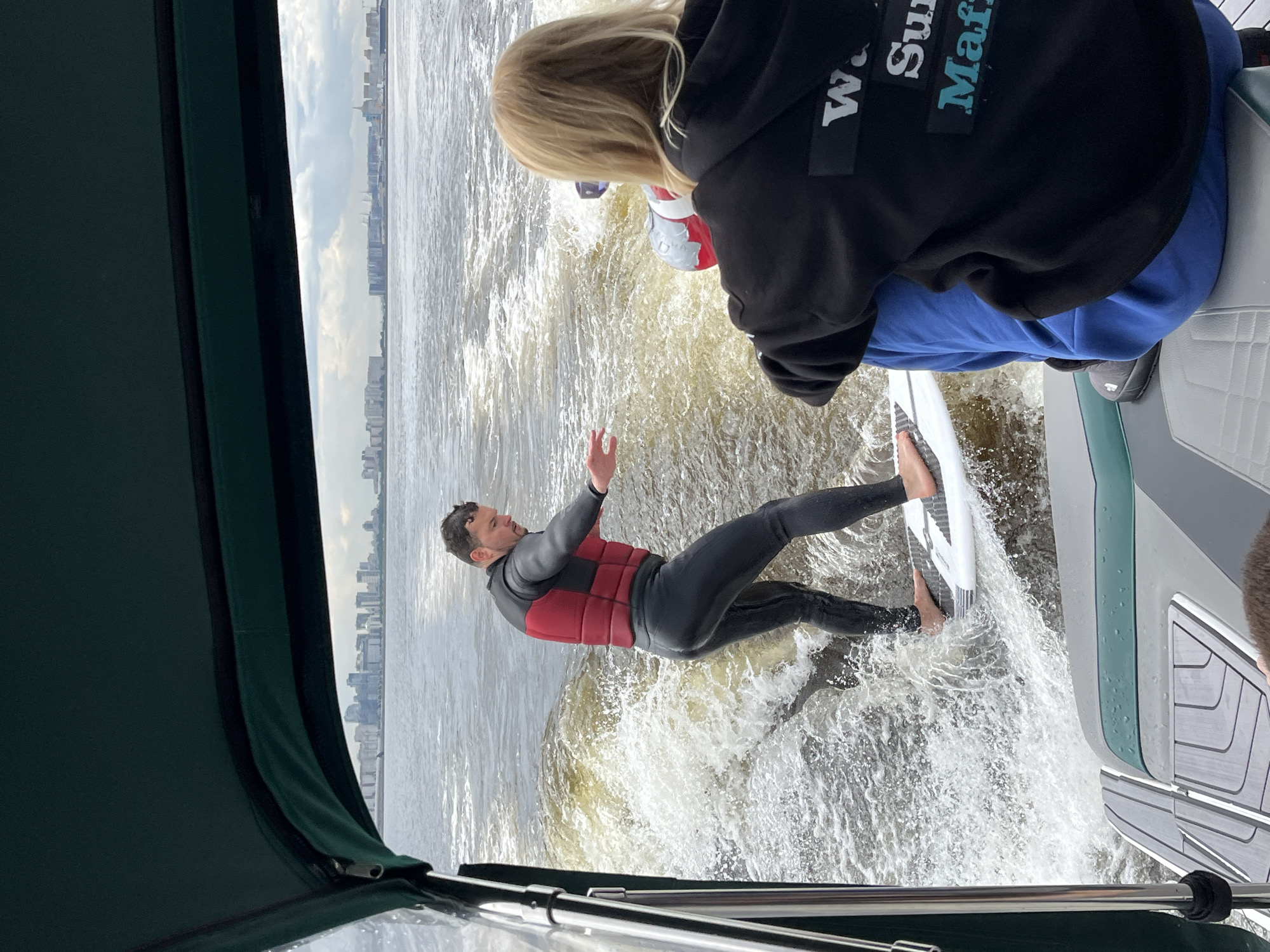 In the foreground, a person with blonde hair wearing a black hoodie with text that reads "W. Surf Mafia" and blue sweatpants is seated facing away from the camera, looking towards the water. To their left, a man in a black wetsuit and a red life vest is wakeboarding. He is on a white board and is being pulled by a boat, creating a large wake. The boat appears to be moving forward. In the background, a city skyline with numerous buildings is visible across the water. The sky is overcast with clouds. The overall scene depicts a recreational activity on the water with a cityscape in the distance, likely within or near the city of Saint Petersburg, Russia.