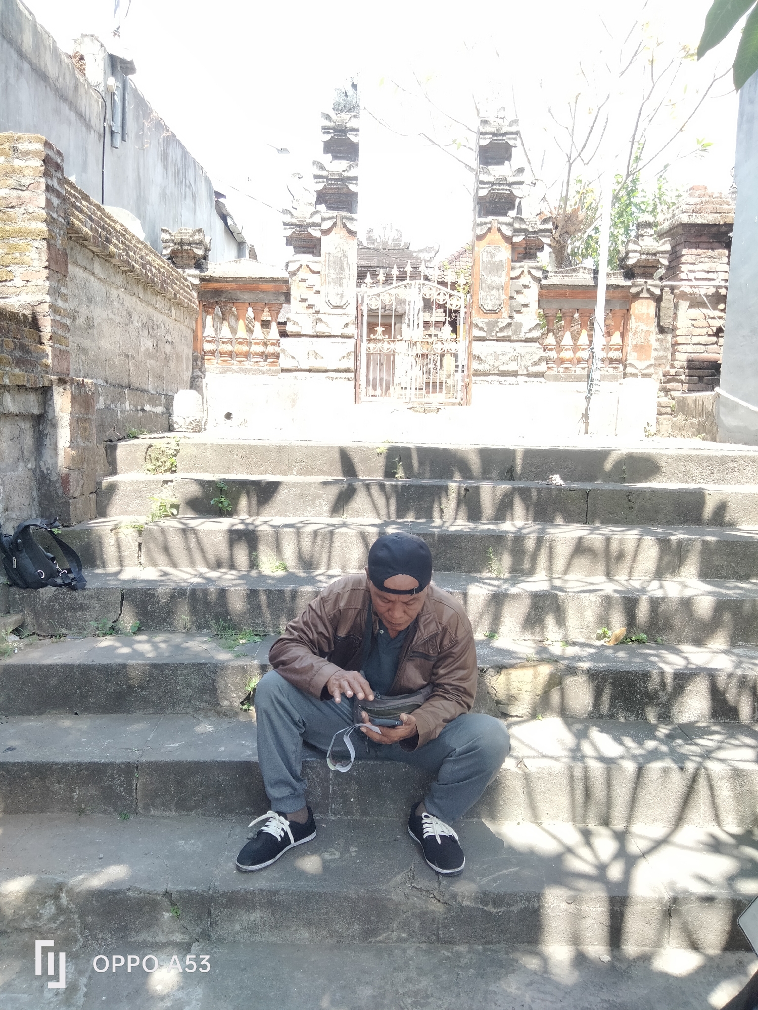 A man wearing a brown jacket, grey pants, and a black baseball cap is sitting on stone steps, looking down at his phone. He is holding a small black bag in his left hand. To his left, a black backpack is placed on a lower step. Behind him, a weathered stone wall and an ornate gate with intricate carvings are visible. The scene is illuminated by sunlight, casting shadows of branches onto the steps. The location appears to be outdoors, possibly in a historical or temple-like setting. The phone's screen is not clearly visible.