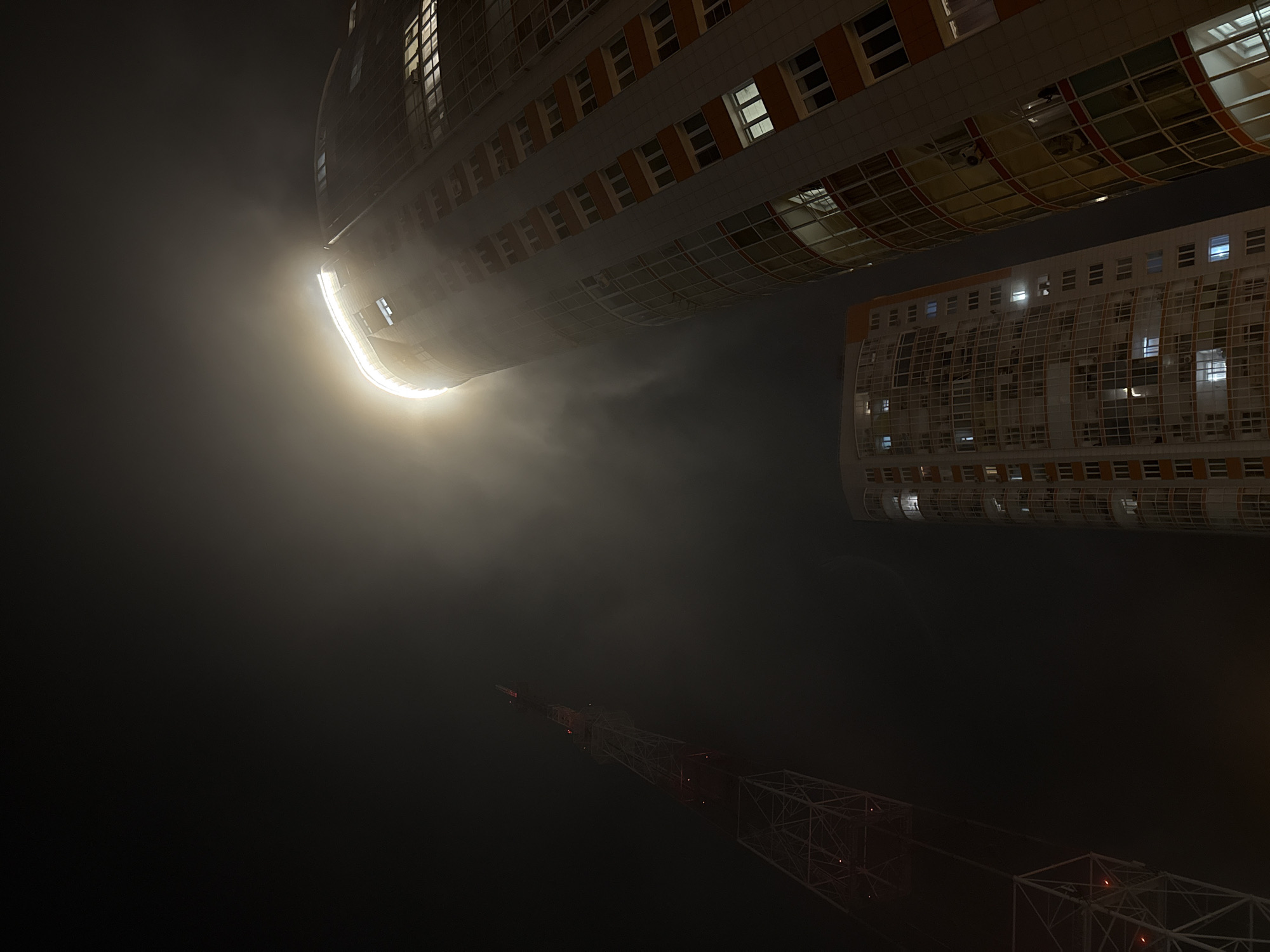 At night in Krasnoyarsk, Russia, a low-angle shot features the curved facades of tall buildings with illuminated windows, likely apartments or offices. A bright light source obscures some details of the top building. A construction crane with red lights is visible against the dark sky. The environment is dark and foggy.