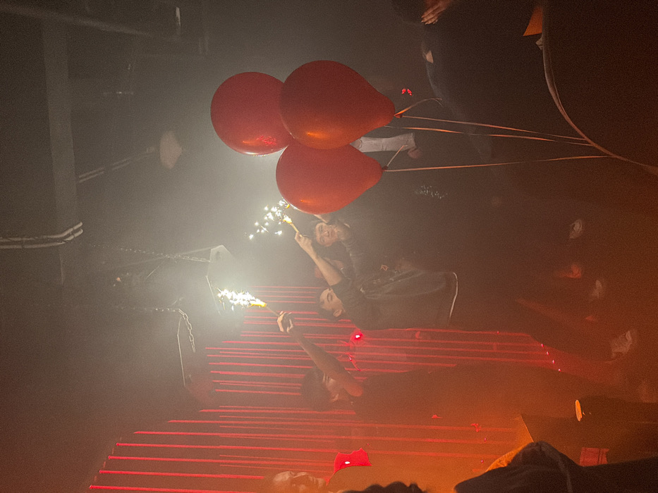 On a staircase illuminated with red lights, two people hold sparklers and red balloons. One individual stands at the top, holding the sparkler aloft. Another person reclines further down the stairs, also with a sparkler. A third person lies at the foot of the stairs. The scene is shrouded in fog and darkness, with other shadowy figures in the background, adding to the enigmatic and dramatic atmosphere.
