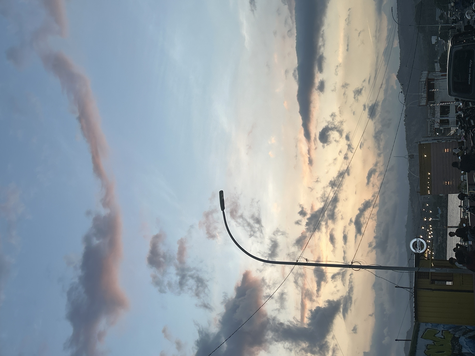 The image depicts a street scene at dusk in Bantaeng, Indonesia. The sky is filled with a mix of gray and pinkish clouds, illuminated by the setting sun, with a pale blue sky above. A tall, curved street lamp is prominent in the foreground, with electrical wires crisscrossing the frame. In the mid-ground, there are several low-rise buildings, one yellow structure with graffiti on its side, and another with a circular sign displaying the word "Hagemony." String lights are visible across the buildings. A body of water, likely a bay or harbor, can be seen behind the buildings. The foreground is populated by a group of people, some seated, and several motorcycles. In the background, hills and mountains form the horizon. The overall atmosphere suggests a lively evening gathering or marketplace by the waterfront.