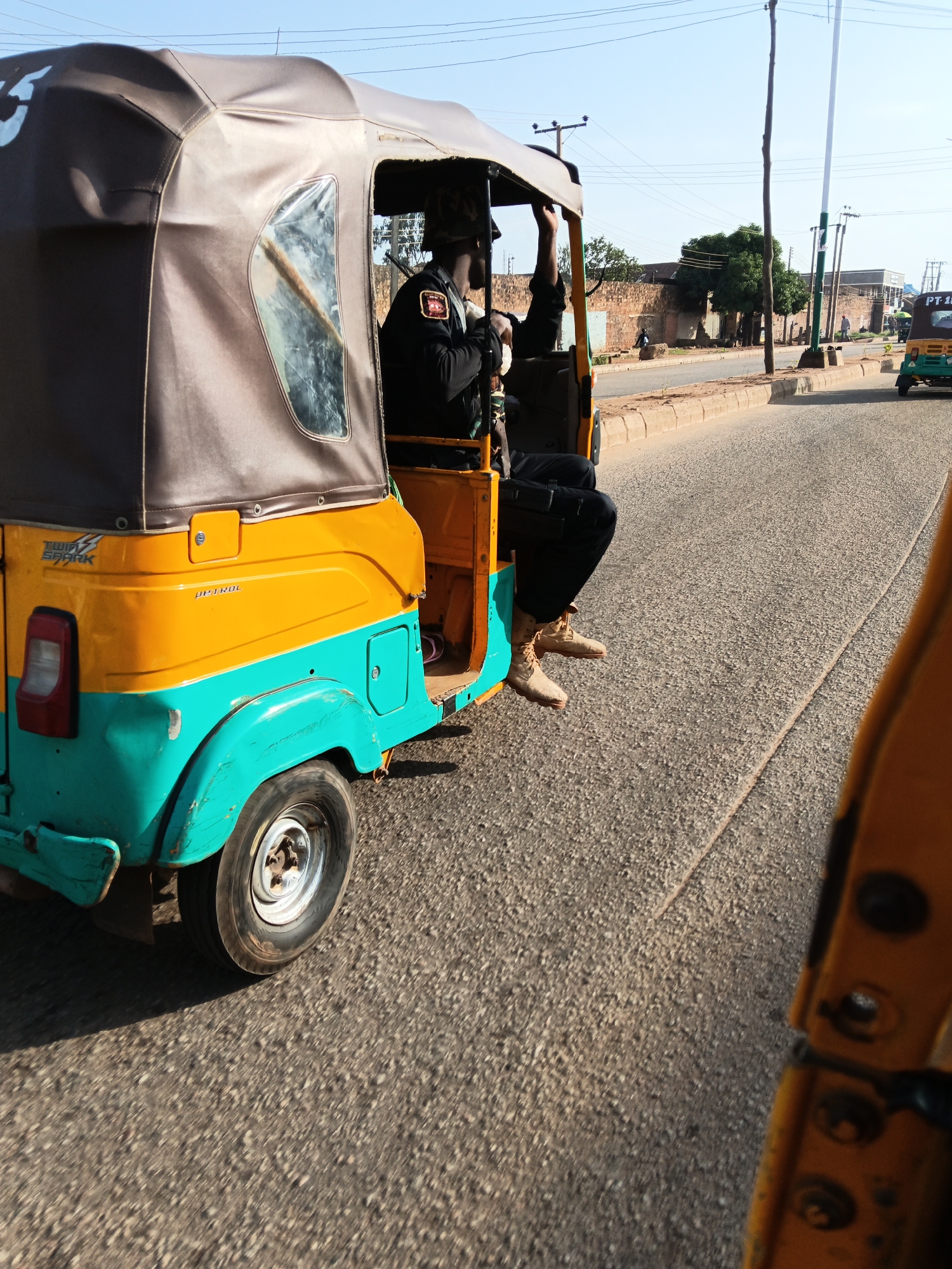 The image depicts a bright, sunny day in what appears to be Bukuru, Nigeria. The primary subject is an auto-rickshaw, a three-wheeled vehicle commonly used as a taxi in many parts of the world. This auto-rickshaw is painted in vibrant yellow and turquoise colors. The driver, dressed in a black uniform with a patch on the sleeve, is visible from the side, holding the steering wheel. The driver is wearing light-colored boots and dark pants.
The auto-rickshaw is in motion on a paved road with a beige concrete curb. The road surface is textured asphalt, suggesting a typical urban or semi-urban environment. In the background, there are buildings with brick walls, some trees, and utility poles with visible overhead wires, indicating an inhabited area. Another auto-rickshaw, this one predominantly green and yellow, is visible further down the road. Some indistinct figures of people can be seen walking in the distance. The sky is clear and blue, suggesting fair weather. The overall impression is that of everyday travel or transit within a community. The perspective suggests the photo was taken from another vehicle moving alongside the auto-rickshaw. There is text visible on the side of the auto-rickshaw in the foreground, reading "TWIN SPARK" and "PETROL". The green auto-rickshaw in the background has "PT 18" visible on its rear.