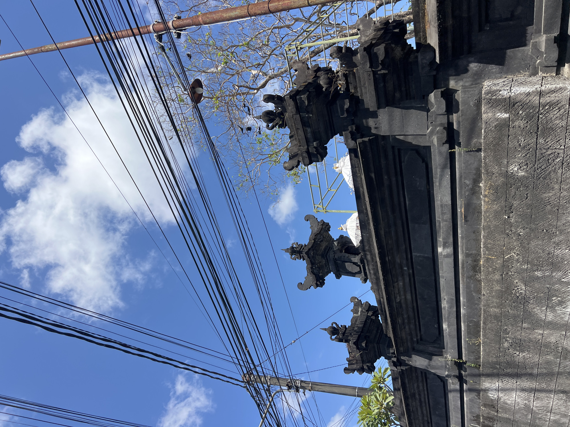 The image shows a traditional Balinese stone wall with ornate carvings, likely part of a temple or a significant building, in Denpasar, Indonesia. The wall is made of dark, weathered stone blocks, with intricate decorative elements on top. Above the wall, a dense network of overhead electrical wires crisscrosses the frame, suspended from utility poles. In the background, a bare tree with intricate branches reaches towards a bright blue sky dotted with white, fluffy clouds. A rusty metal lamppost is also visible, further adding to the urban streetscape. The overall scene depicts a blend of traditional architecture and the ubiquitous infrastructure of a modern city, under a clear, sunny sky.