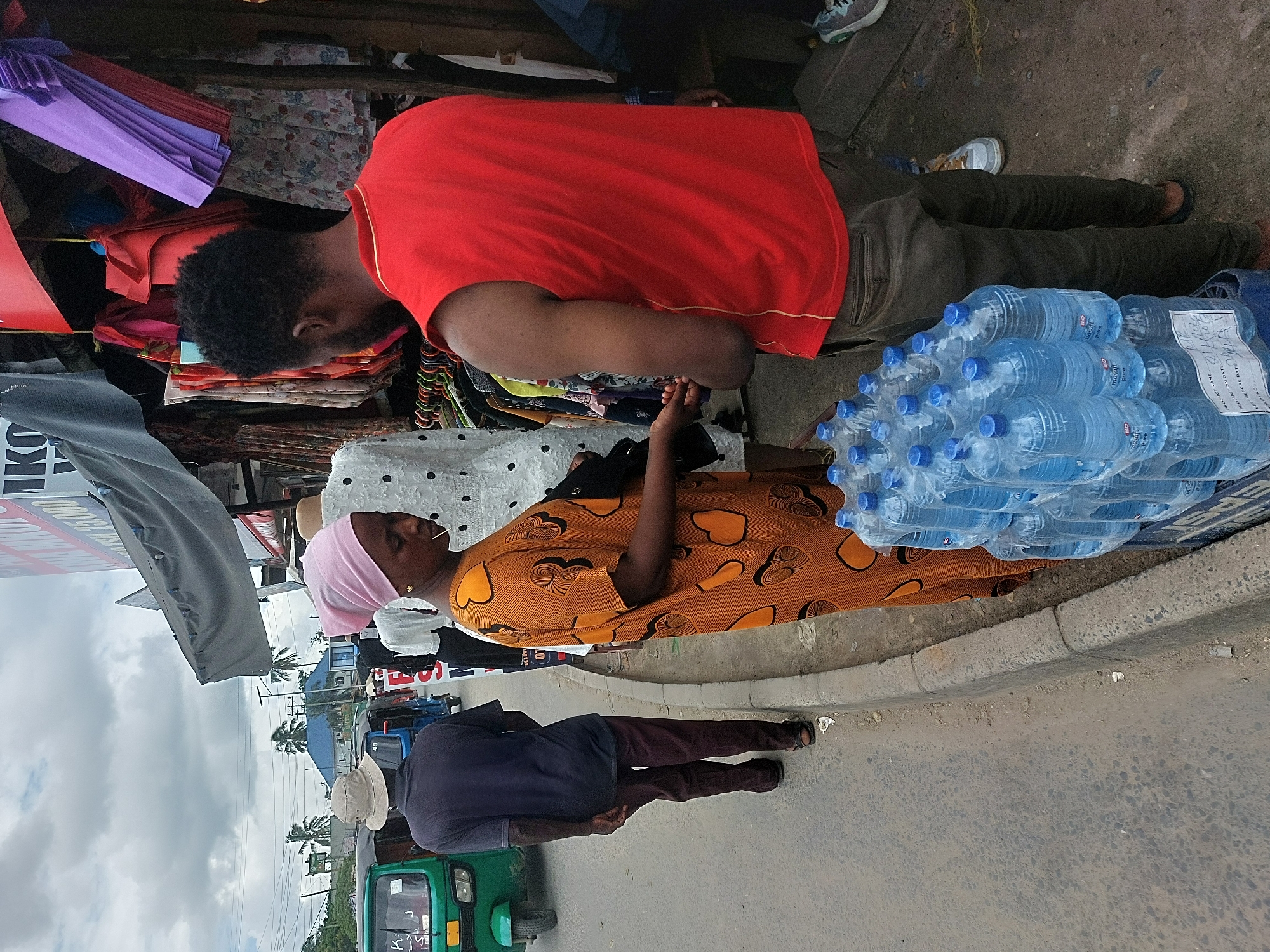 The scene depicts a street in Dar es-Salaam, Tanzania. In the foreground, an African woman with a pink headscarf is wearing a brown and orange patterned dress and appears to be selling bottled water stacked on a table or cart along the roadside. A man in a red shirt is seen browsing through clothing or goods displayed on the same cart. Another man in a dark shirt and a light-colored hat stands near a green tuk-tuk or auto-rickshaw on the road. The street is partially lined with various buildings and the sky is overcast.