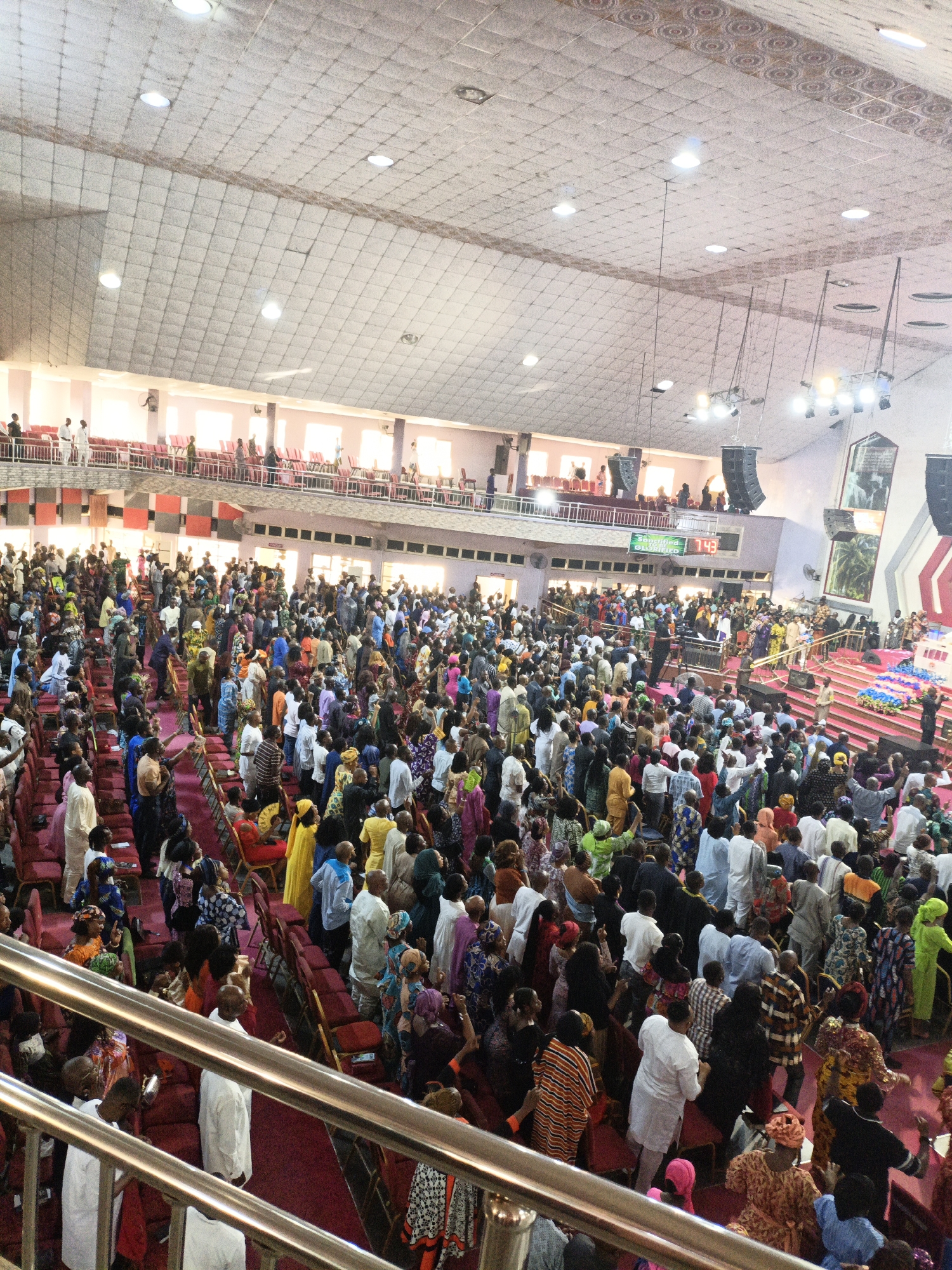 A large group of people is gathered in an expansive indoor space, seemingly a church or auditorium, in Sabon Gari West, Nigeria. The attendees fill the seating area, with many standing. The hall is large, with a balcony level visible in the background where more individuals are standing. A stage is positioned at the far end of the hall, indicating a focal point for speeches, performances, or religious services. The ceiling is high and expansive with various ceiling lights, suggesting a modern structure. The crowd is diverse in clothing, with a mix of colors and patterns. The environment suggests a large gathering for a special event or regular service.