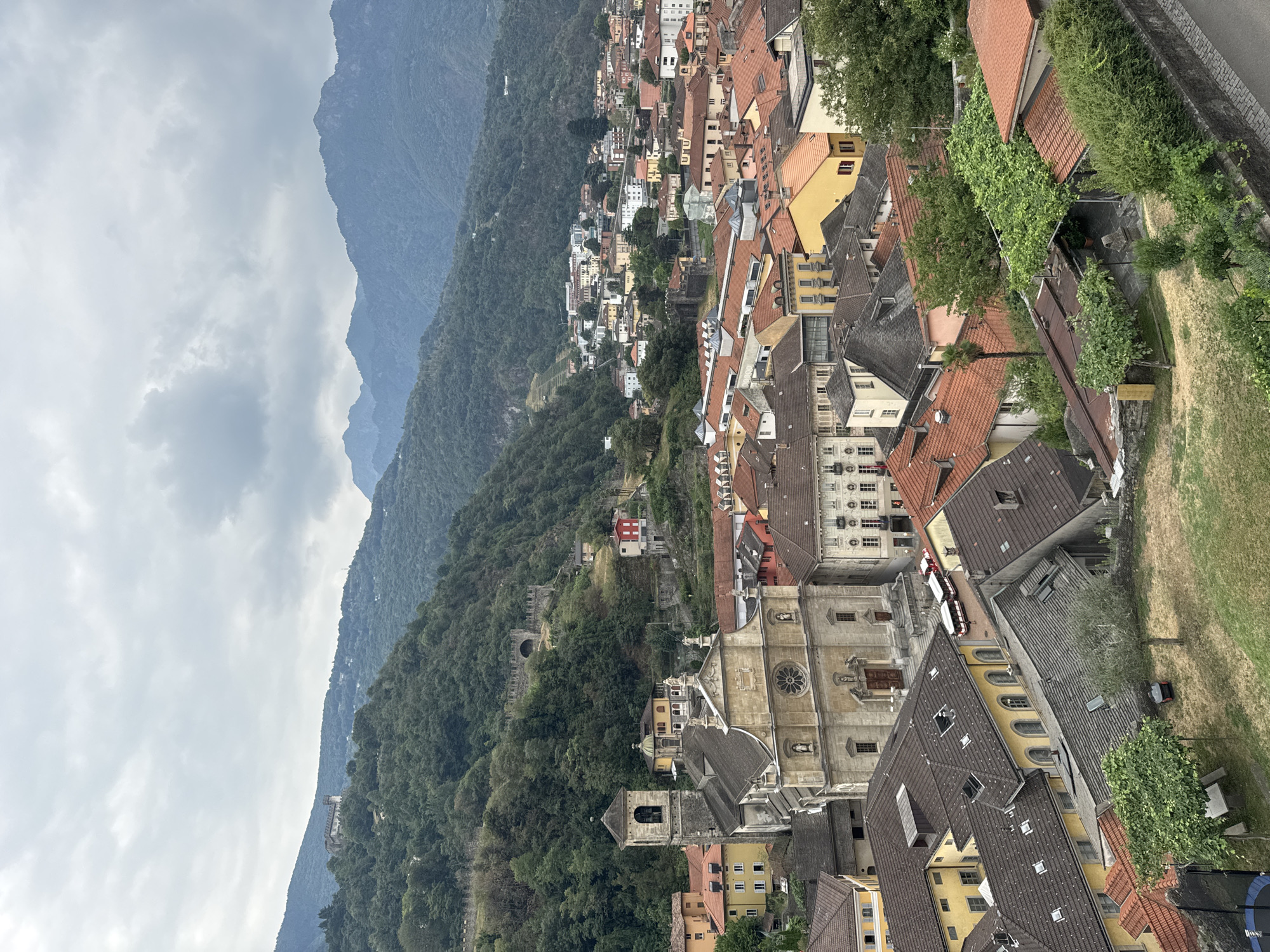 This is a high-angle shot of a town nestled in a valley, with mountains in the background. The foreground features a prominent church with a bell tower, surrounded by buildings with terracotta roofs. The architecture suggests a European setting, likely historic. In the middle ground, more buildings are visible, and the slopes behind them are covered in dense green trees. The sky is overcast with gray clouds. Further in the background, the mountains rise, some with visible fortifications or ruins on their slopes. The town appears to be Bellinzona, Switzerland, known for its medieval castles. Two red and white buses are parked on a street near the church. The overall scene depicts a charming, historical town integrated with its natural mountainous environment.