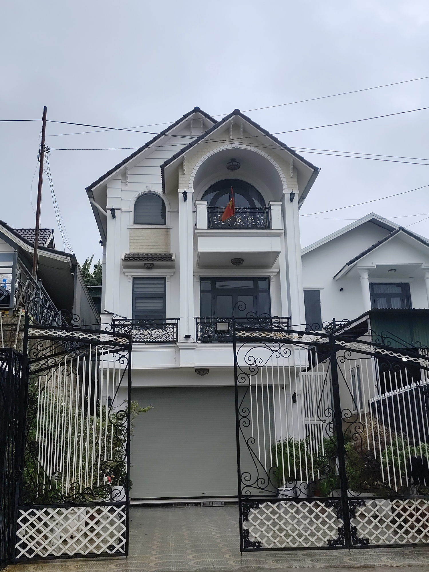 The image depicts a multi-story white house with a dark tiled roof, likely located in Lạc Dương, Vietnam. The house features an arched balcony on the second floor, adorned with an ornate black railing and a Vietnamese flag. The front of the property is secured by a tall black metal gate with decorative scrollwork and white vertical bars. A grey roller shutter garage door is visible behind the gate. The ground is paved with patterned tiles. The sky is overcast, and a utility pole with electrical wires runs along the left side of the frame. Adjacent to the main house, another white building with a similar roofline is partially visible to the right, and a section of a neighboring house with a dark roof and stairs is seen to the left. Lush green foliage is visible in planters and around the property.