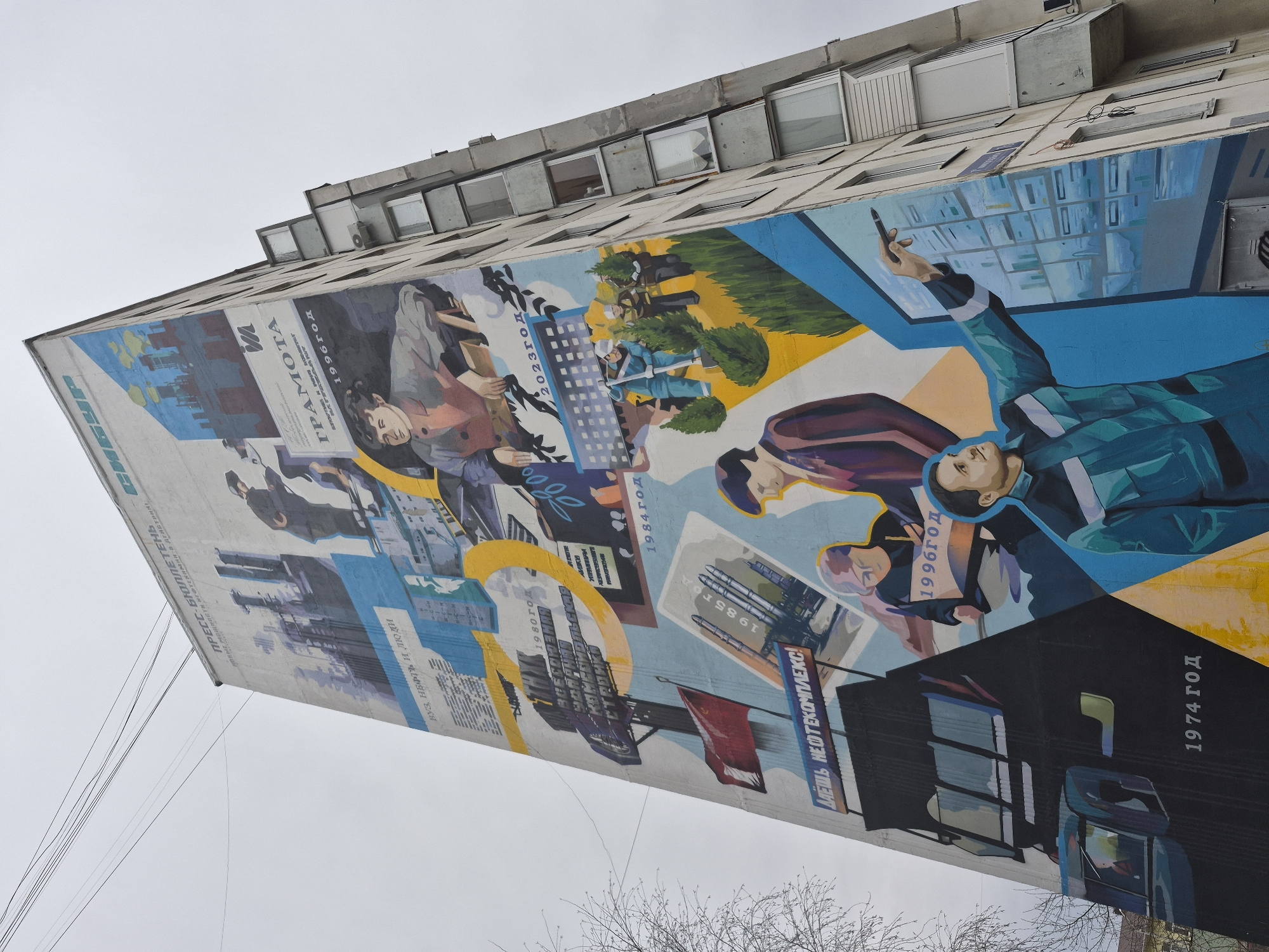A large mural, painted on the side of a tall, grey concrete building in Tobolsk, Russia, is captured from a low angle under an overcast sky. The mural depicts a chronological timeline of the city's industrial development, particularly related to the petrochemical industry, spanning from 1974 to 2023.
The artwork begins at the bottom right with "1974 ГОД" (1974 Year), featuring dark industrial elements and the rallying cry "ДАЁШЬ НЕФТЕКОМПЛЕКС!" (Give us the petrochemical complex!). Moving upwards and across, "1980 ГОД" (1980 Year) shows industrial buildings and the text "ВУЗ, НЕФТЬ И ЛЮДИ" (University, Oil, and People). Further along, "1984 ГОД" and "1985 ГОД" depict rocket-like structures, symbolizing technological advancements.
The "1996 ГОД" (1996 Year) section portrays people, including a woman holding a document labeled "ГРАМОТА" (Certificate/Diploma), indicating achievement. The most recent section, "2023 ГОД" (2023 Year), illustrates a modern scene with professionals; one man points towards a grid-like digital display, while another group in blue uniforms appears to be planting trees, representing urban and environmental development.
Additional text visible along the far left edge includes "ПРЕСС-БЮЛЛЕТЕНЬ" (Press Bulletin) and "МОЩНОСТИ НЕФТЕХИМИИ В ДЕЙСТВИИ!" (Power of Petrochemistry in Action!). The overall scene conveys a narrative of continuous progress and growth. Bare tree branches are visible at the bottom, and thin wires stretch from the building against the dull, grey sky. The time of day is daytime, but the weather is cloudy, lending a subdued light to the scene.