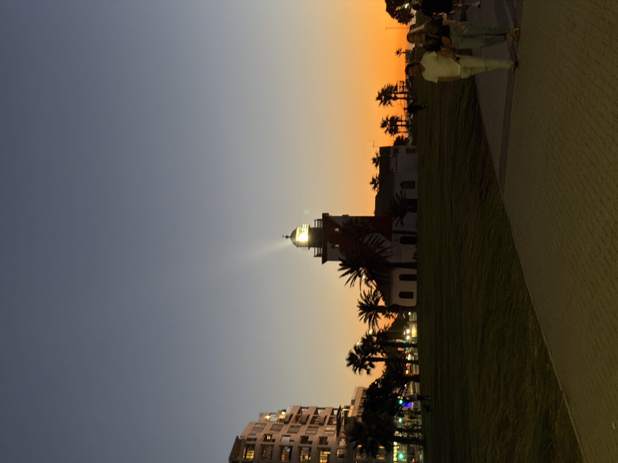 The photograph captures a scene at dusk in Cape Town, South Africa, featuring the Mouille Point Lighthouse. The lighthouse, with its illuminated beacon, is centrally positioned amidst palm trees and adjacent buildings. The sky transitions from a dark blue to an orange hue, indicating the setting sun. A building with lit windows sits below the lighthouse. On the right side of the image, a paved walkway is visible with people standing or walking. Green grass separates the walkway from the lighthouse.