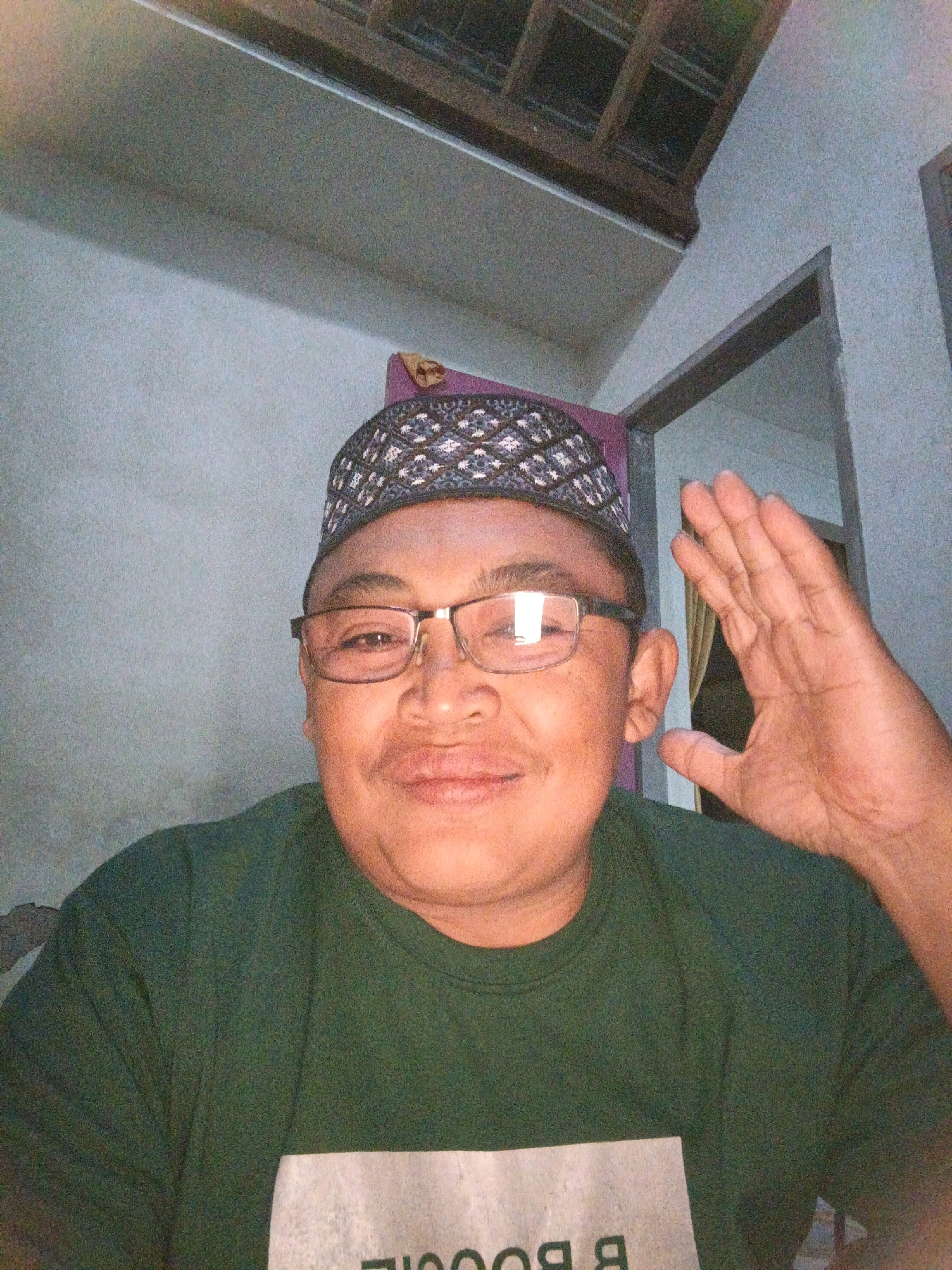 A medium shot captured from a slightly low angle shows a person wearing a dark green t-shirt and a patterned skullcap, looking at the camera. The individual is wearing glasses with dark frames. Their right hand is raised and open, palm facing the camera, as if waving. The background consists of a plain, light-colored wall and a ceiling with exposed wooden beams. The overall lighting is dim, suggesting an indoor setting. The specific location is identified as Ketegan, Indonesia.