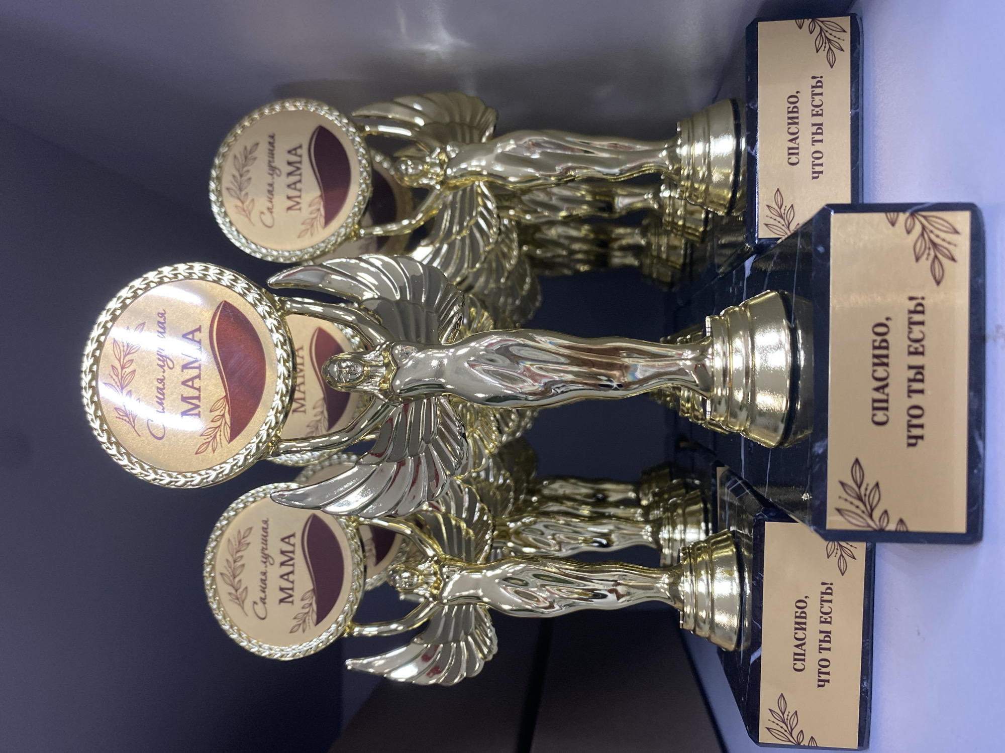 The image depicts a close-up of three identical gold-colored trophies, each featuring a winged female figure. Each trophy has a round medallion with the inscription "Самая лучшая МАМА," which translates to "The best MOM" in Russian. The base of each trophy has a black marble-like finish with a gold plate inscribed with "СПАСИБО, ЧТО ТЫ ЕСТЬ!" meaning "THANK YOU FOR EXISTING!" in Russian. It is most likely that the location of this picture is Chaikovsky, Russia, since that is the region that the Russian language is most commonly spoken.