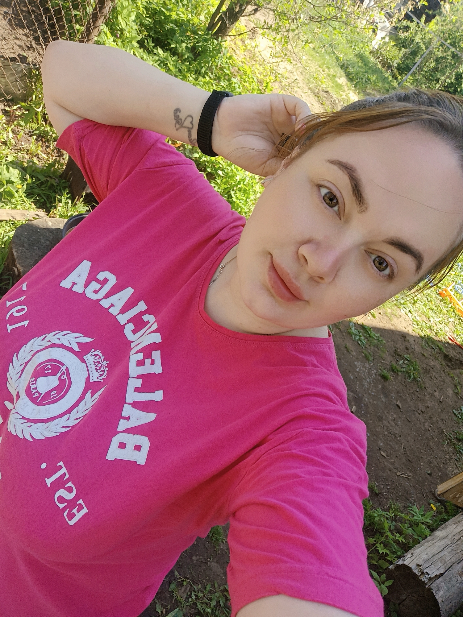 A woman is taking a selfie outdoors. She is wearing a bright pink T-shirt with white lettering and a logo. Her hair is pulled back. She is holding one arm up near her head. The background shows green foliage and glimpses of a garden or rural setting. The location appears to be in a natural or semi-rural area. The specific location within Chaikovsky, Russia, cannot be determined from the image alone.