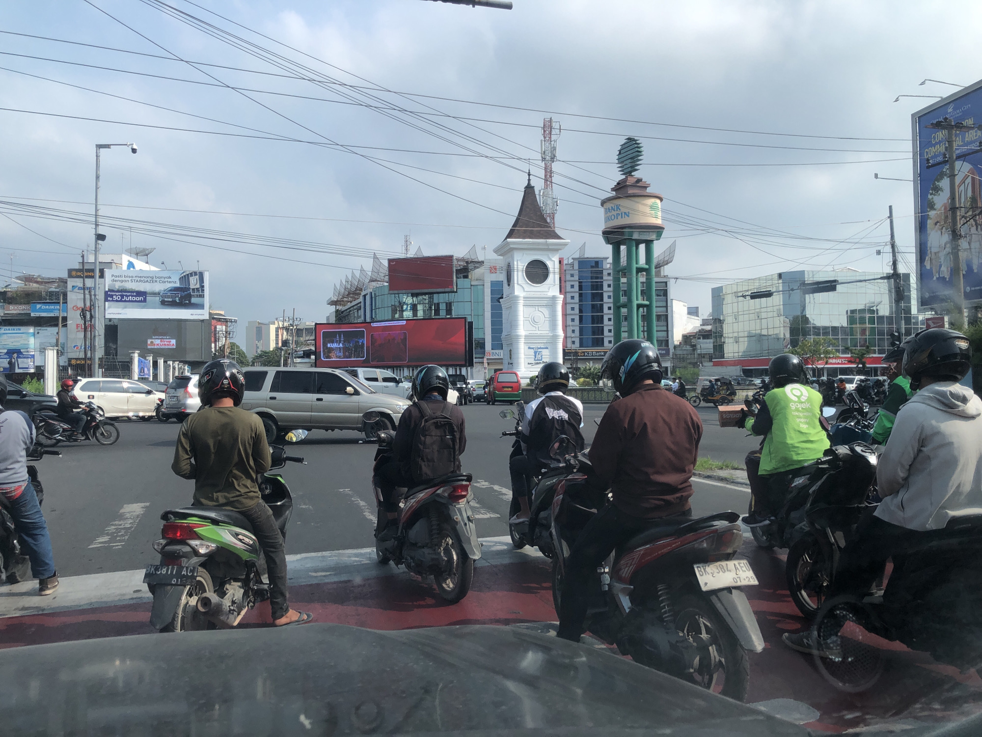 In a city intersection in Medan, Indonesia, multiple motorcyclists are stopped at a traffic light. The motorcyclists are dressed in casual clothing, with several wearing helmets. One motorcyclist in the foreground is wearing a dark green long-sleeved shirt and black pants, with a green and black motorcycle. Another motorcyclist, wearing a white hoodie and black pants, is on a black motorcycle. A third motorcyclist wears a dark brown jacket and black pants, with a red and black motorcycle. Several other motorcyclists are visible in the background, some with helmets and backpacks.
The environment is a busy city street with several buildings and billboards. A tall water tower with the logo "BANK KOPIN" is visible in the background, along with a clock tower. Traffic lights are present, and the road is marked with white and red lines. Several cars are also present in the intersection, indicating a moderate amount of traffic. The sky is overcast, suggesting a cloudy day. The scene appears to be a typical urban setting in Indonesia.