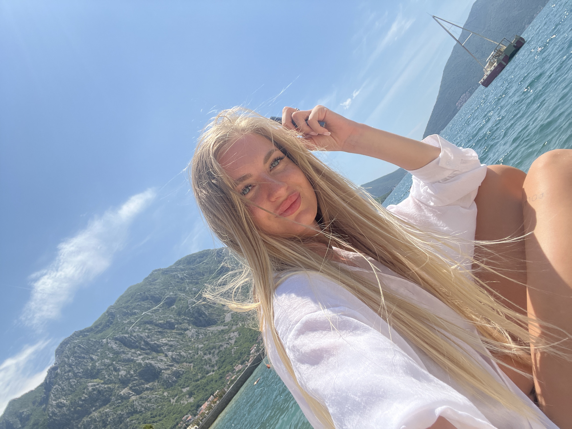 The media file shows a young woman with long blonde hair taking a selfie outdoors. She is wearing a white, loose-fitting garment, possibly a shirt or cover-up. Her left hand is raised, touching her hair, and she is smiling at the camera. The background features a bright blue sky with scattered clouds, and a body of water that appears to be a bay or sea. To the left, a lush, green mountainside slopes down towards the water, with some buildings visible near its base, suggesting a coastal town. On the right, a sailboat with a tall mast is visible on the water. The scene suggests a sunny day, likely during daylight hours, and the overall mood is relaxed and happy. The location cues, particularly the mountains and the water, are consistent with a Mediterranean coastal environment. No visible text is present in the media file.