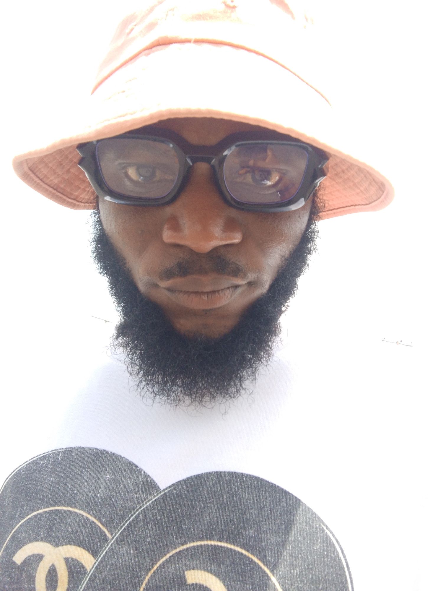 A close-up photograph shows a person with dark skin wearing a light orange bucket hat and black-framed eyeglasses. The individual has a thick, dark beard. The person is wearing a white t-shirt with two overlapping, black circular designs on it, each with a gold Chanel logo. The location is unknown.