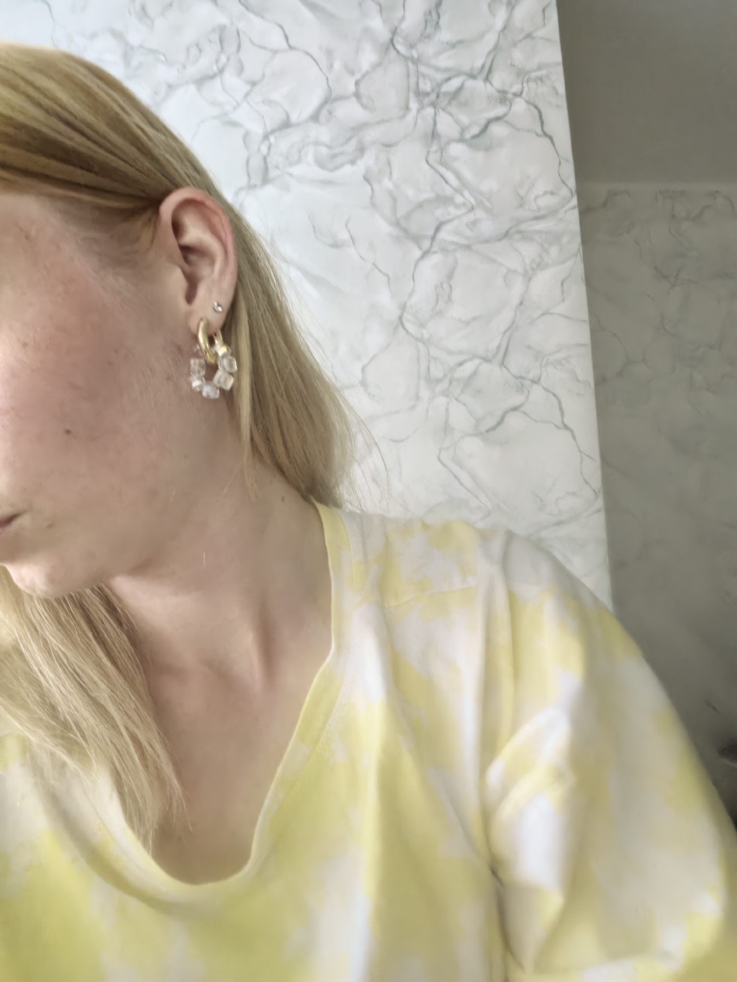 A woman with blonde hair wears a yellow and white tie-dye shirt and gold hoop earrings with clear, geometric charms. She has a piercing in her ear. The background is a white wall with grey marble-like patterns. The location and city of Sterlitamak, Russia, cannot be determined from the image.