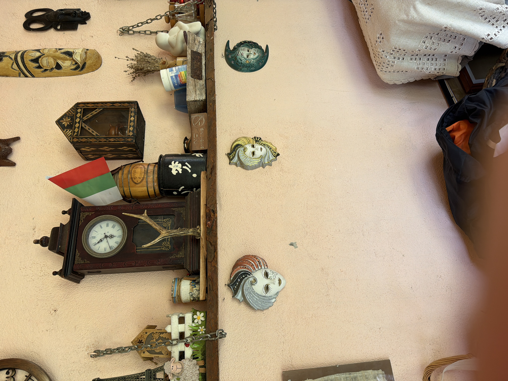 This is a detailed, forensic, matter-of-fact description of a scene in the city of Lobosh, Bulgaria. The image captures a wall adorned with various decorative objects and shelves. On the wall, there are at least three decorative masks, two of which are prominently displayed and appear to be ceramic or plaster. One mask, on the left, is white with silver and reddish-brown accents, featuring a stylized hairstyle. Another mask, slightly to the right and further down, is a crescent moon shape in a teal color, with a face depicted within it. A third mask, between the first two but higher up and to the right, is also white with gold and black accents. Above the masks, a wooden shelf is mounted on the wall. On this shelf, several items are arranged. To the left, there is a dark cylindrical object with a white floral design. Next to it is a small wooden structure with a glass front, possibly a display cabinet, containing what appears to be a small wooden object. To the right of this cabinet, a wooden barrel with a Bulgarian flag (white, green, red) is placed. Further to the right on the shelf, a bundle of dried lavender or similar herbs is tied with twine. Beside the herbs, there is a white plastic container with a blue lid, possibly medication or a cosmetic product, and a white ceramic figurine of a person. To the left of the shelf, a large, dark wooden mantel clock with a Roman numeral clock face is mounted on the wall. A pair of deer antlers is attached to the front of the clock. Below the clock and to its left, a decorative chain hangs, connected to a small white fence with grass and flowers. An Eiffel Tower replica is also visible on the left side, partially obscured by the fence. Further to the right on the wall, above the shelf, are two carved wooden masks. One is a long, light-colored mask with black and yellow intricate designs. Another, to its right and slightly lower, is a smaller black mask. The wall itself is textured and painted a light peachy-pink color. The overall impression is one of a decorated interior wall with a collection of eclectic decorative items. The bottom of the image shows a portion of a person's hand, and below that, a folded black garment with an orange interior is visible, along