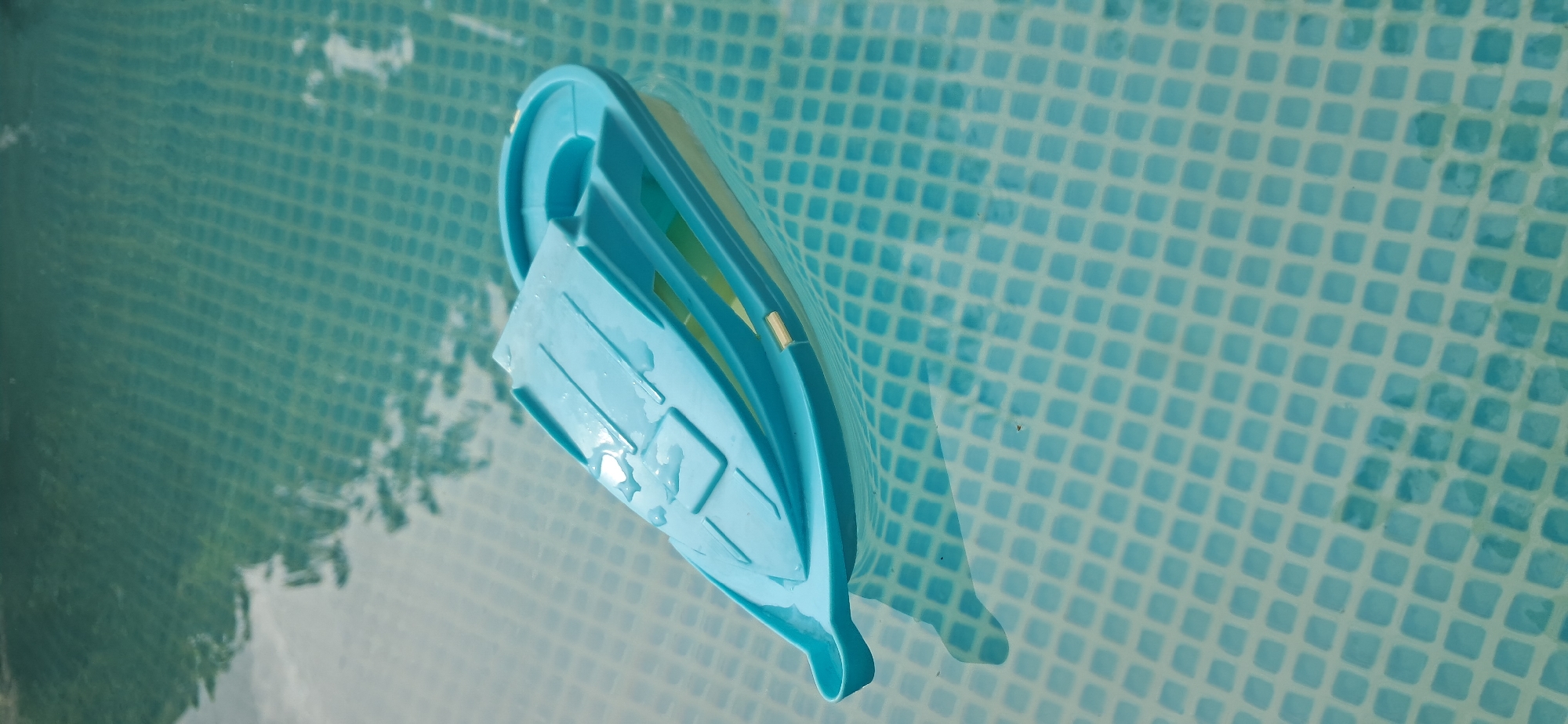 A light blue plastic toy boat floats on the surface of a swimming pool. The boat is partially filled with water and has some water droplets on its deck. The pool water is clear and has a tiled bottom, visible as a grid pattern. Reflections of trees and sky are visible on the water's surface. The image does not contain any information that would allow for its location to be determined in Kryvyi Rih, Ukraine, nor does it depict any people, actions, or interactions.