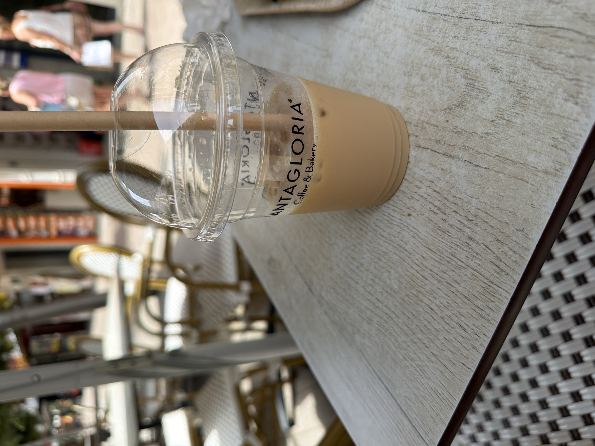 An iced coffee drink in a clear plastic cup with a domed lid and a brown paper straw sits on a light-colored wooden table. The cup has the "SANTAGLORIA Coffee & Bakery" logo printed on it in black. The drink is a light brown color and contains ice cubes. In the background, out of focus, are tables and chairs of an outdoor cafe, with several people walking in the distance. The setting appears to be a pedestrian street or plaza in Benidorm, Spain, characterized by the style of outdoor seating and the bright, sunny lighting. The table has a textured surface with visible wood grain. The lower part of the image shows the woven texture of a placemat or the edge of the table.