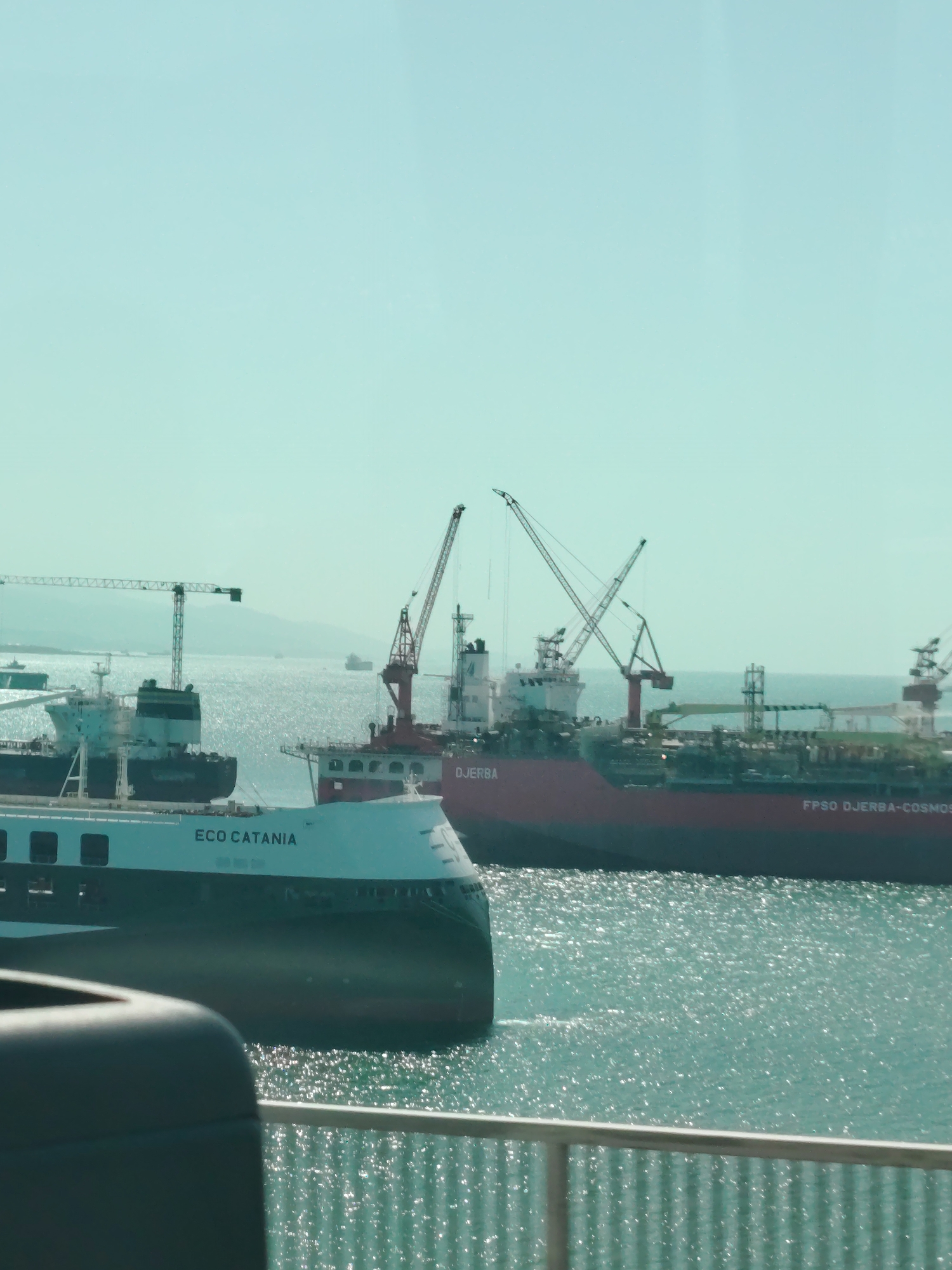 The image captures a maritime scene in a harbor or shipyard, likely in Altınova, Turkey, under a bright, clear sky. In the immediate foreground, a dark, indistinct object and a grey railing suggest the photo was taken from behind a barrier or from within a vehicle.
The midground is dominated by two large vessels. To the left, a white and dark green ship, possibly a cargo vessel or ferry, is prominent, with the name "ECO CATANIA" clearly visible on its side. Its bow is angled towards the viewer. To the right, a substantial red industrial vessel is seen, featuring multiple cranes on its deck. The name "DJERBA" is visible on its side, and further along the hull, "FPSO DJERBA-COSMOS" can be read, indicating it is a Floating Production Storage and Offloading unit.
The calm water in the harbor sparkles intensely, reflecting bright sunlight across its surface, suggesting a sunny day with good visibility. In the background, beyond these primary vessels, other ships, including some with cranes, are partially visible, indicating a busy port or shipbuilding area. Distant land or mountains appear hazy on the horizon under the pale blue sky. No specific human activity is discernible, but the presence of these large industrial ships implies ongoing maritime operations, such as docking, loading, or maintenance.