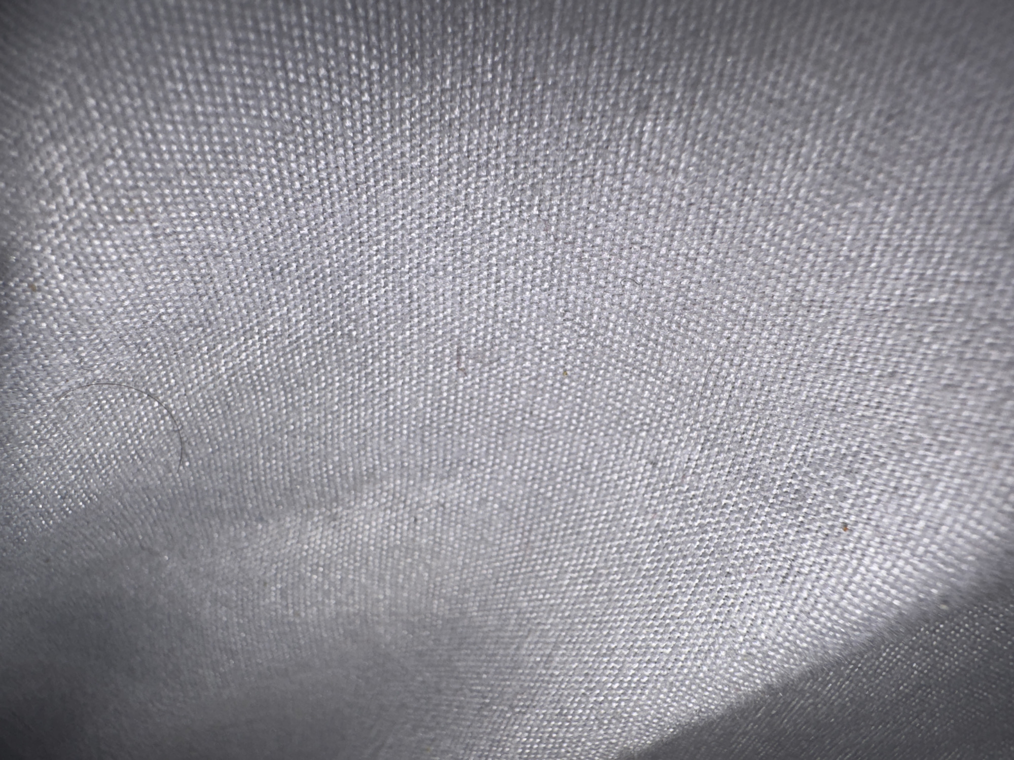 The image displays a close-up, textured view of a grey fabric, possibly linen or a similar textile, with a visible weave pattern. The lighting creates subtle highlights and shadows across the surface, suggesting a slightly uneven or draped texture. There are no discernible people, objects, or specific environmental features that would indicate a location within Al Buraimi Province, Oman. The image is purely a detail shot of fabric.