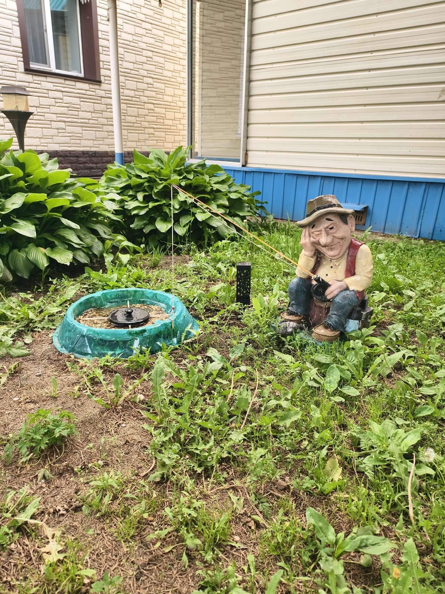 The image depicts an outdoor scene in Belogorsk, Russia, featuring a garden area with a focus on a decorative garden gnome. The gnome, sculpted in the likeness of an elderly fisherman, is seated on a stool and appears to be fishing, with a fishing rod extending upwards. It is wearing a hat, a vest over a light-colored shirt, and blue pants. The gnome is positioned amidst lush green grass and various types of vegetation, including some broad-leafed plants. To the left of the gnome, there is a circular water feature, possibly a bird bath or a small pond, with a black object at its center. In the background, the facade of a building is visible, constructed with beige, horizontally oriented siding. The building has a window with brown trim and a white downspout. A blue trim runs along the base of the building. The overall environment suggests a residential backyard or garden.