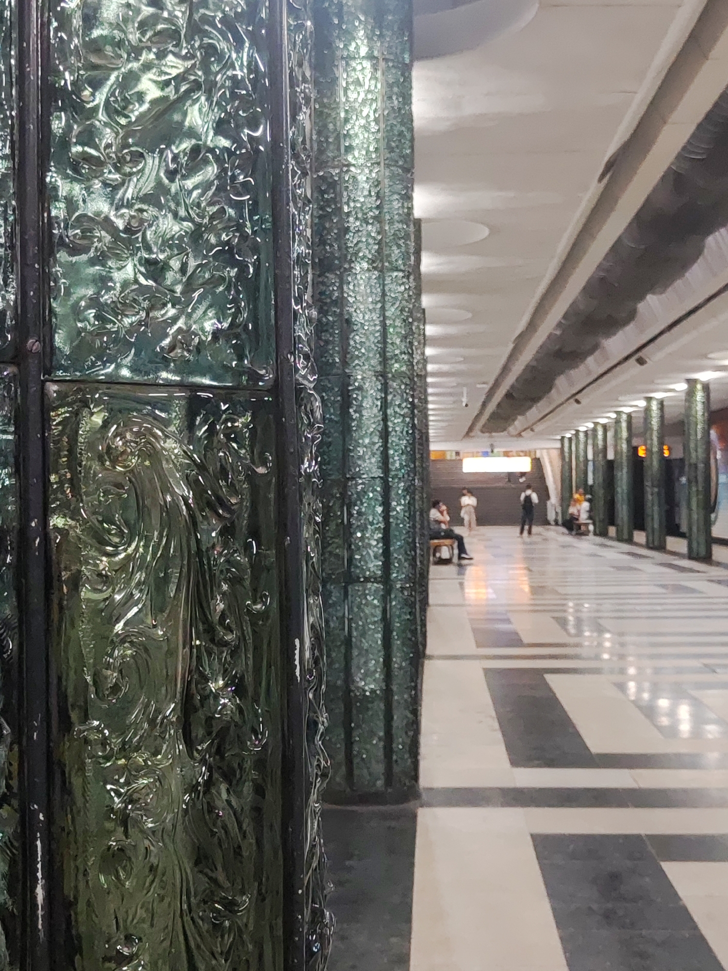 The image depicts a subway station in Tashkent, Uzbekistan, likely "Paxtakor" station, based on its distinctive architectural features. In the foreground, a pillar covered in textured green glass panels with ornate floral motifs dominates the left side of the frame. These panels appear to be made of thick, molded glass, reflecting the ambient light and creating a shimmering effect.
The background reveals the long, linear design of the subway platform. Several identical pillars, also adorned with the same green glass, line the right side of the platform, receding into the distance. These pillars are spaced regularly, creating a rhythmic pattern. The ceiling is high and features recessed lighting and circular patterns.
The floor is paved with large, geometric tiles in a contrasting pattern of light and dark grey or black, and white or cream. The polished surface of the floor reflects the overhead lights and the pillars.
In the middle ground and background, several individuals are visible. On the left side, a person is seated on a bench. Further down the platform, several more individuals are either standing or seated on benches, waiting. One person in the center is standing and appears to be looking towards the stairs or escalator in the distance. The overall atmosphere is one of quiet anticipation, typical of a public transit space. The presence of a CCTV camera mounted on one of the pillars suggests security measures. The lighting is bright and functional, illuminating the spacious area.