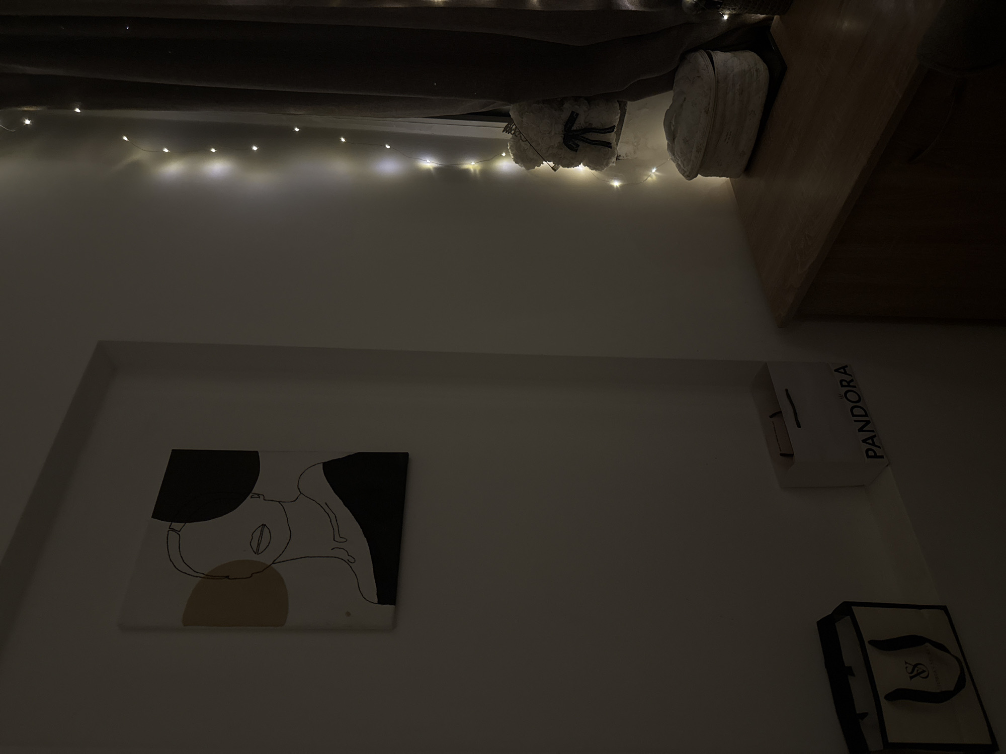 A low-angle, indoor shot depicts a bedroom scene. A painting with abstract figures in black, white, and ochre is mounted on a wall. Above it, fairy lights are strung across the wall's upper portion, below a bed frame. On a nearby nightstand, a pair of fluffy slippers are visible. Two branded shopping bags, one from Pandora and another, partially visible, with a "VS" logo, are positioned on the wall to the right. Sofia, Bulgaria, is not discernible from this image.