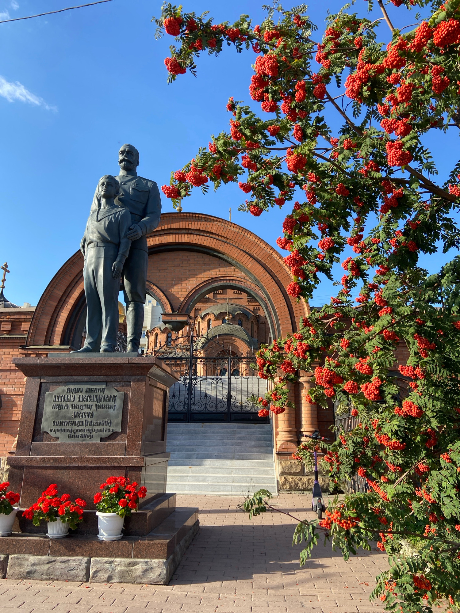 The image depicts a statue of a man and a child in front of a brick building with an arched entrance and domes, situated in Novosibirsk, Russia. The statue is made of dark metal and stands on a stone pedestal with a bronze plaque. The man, wearing a military uniform, stands behind and to the right of the child, with his arm around the child's shoulder. The child, dressed in a sailor's uniform, looks to the left.
The building behind the statue appears to be a church, with a cross visible on its roof. The entrance is large and arched, with ornate metal gates. The area in front of the building is paved with bricks and features a set of stairs leading up to the entrance.
A tree laden with bright red berries, likely rowanberries, is in the foreground on the right side of the image, partially obscuring the view of the building. Red flowers in white pots are placed at the base of the statue.
The sky is clear and blue, suggesting a sunny day. The overall scene suggests a memorial or a historical site within a city setting. The plaque on the pedestal likely contains information about the individuals depicted in the statue and their significance.