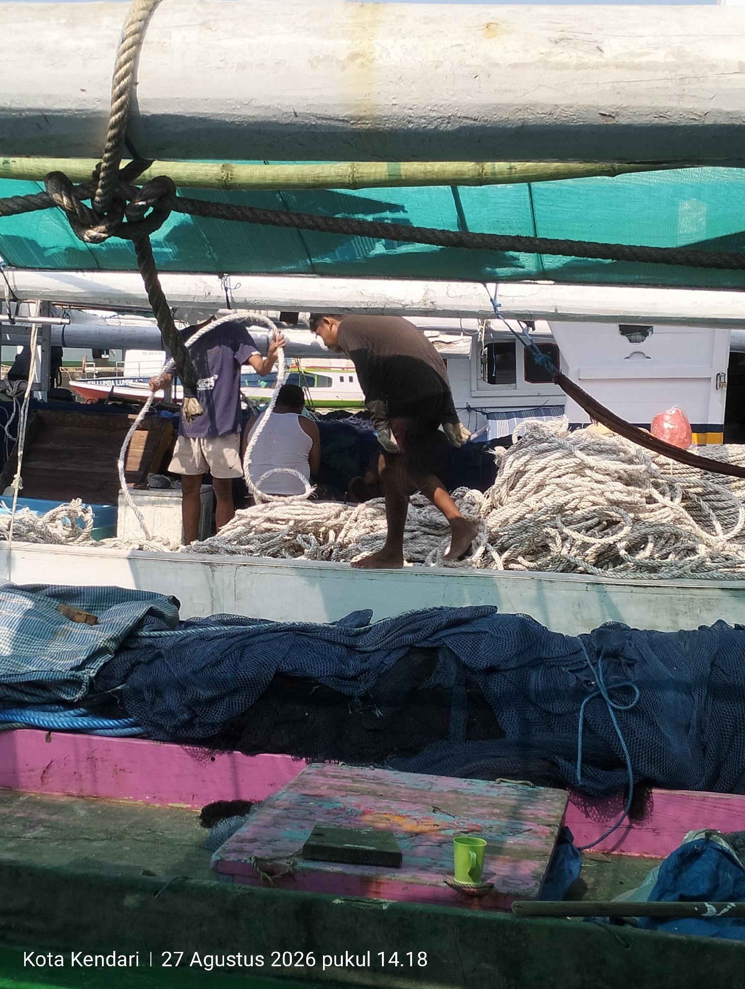 This image shows a group of men working on a boat in Kendari, Indonesia, on August 27, 2026, at 2:18 PM. The scene is on deck, with various ropes and fishing nets visible. In the foreground, there are stacks of coiled ropes and dark blue fishing nets. A pink-painted wooden structure runs along the edge of the boat, with a green cup resting on it. In the mid-ground, three men are present. One man, wearing a dark blue t-shirt and light-colored shorts, is holding a thick white rope. Another man, wearing a white tank top, is also holding a rope. A third man, wearing a dark shirt, is in the process of moving or working with the ropes. Behind them, the white superstructure of another vessel is visible, along with a red bag. The overall environment suggests a working harbor or port.