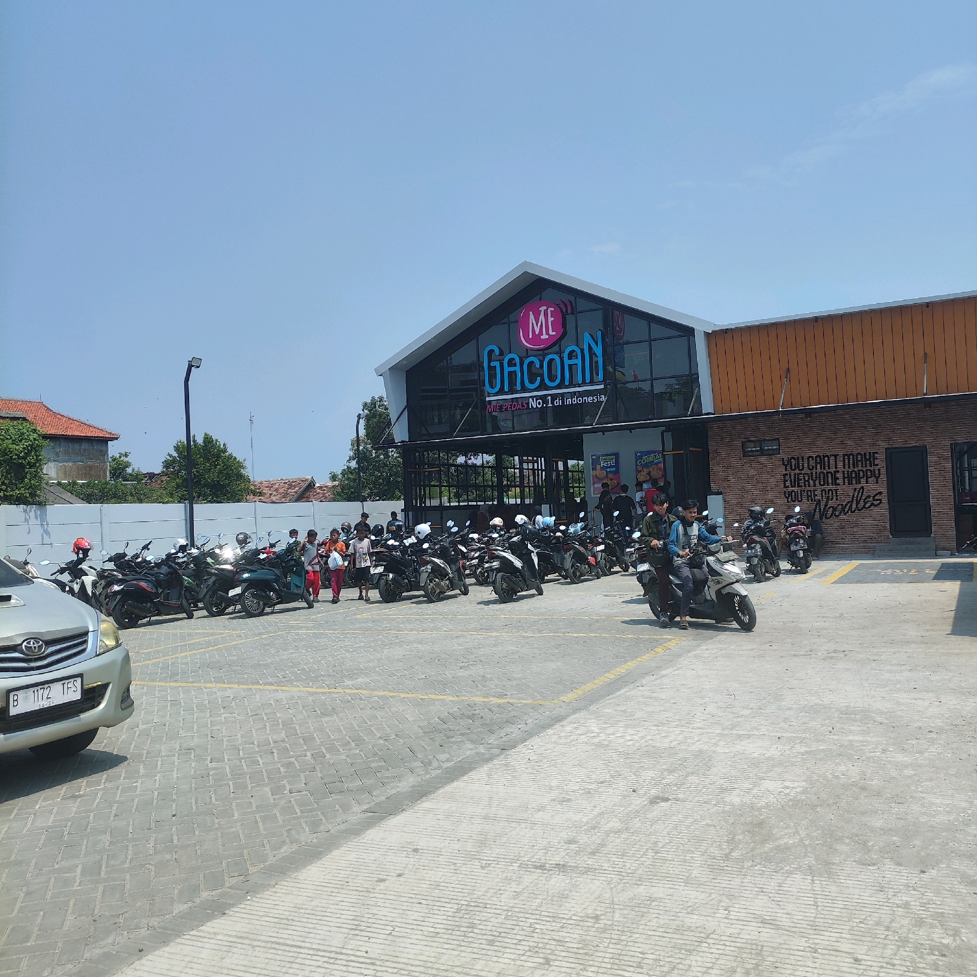 This is a wide shot of a restaurant parking lot on a clear day. The restaurant, "ME Gacoan," has a modern design with large windows and a prominent neon sign. In front of the restaurant, a large number of motorcycles are parked. Several people are walking and standing near the motorcycles and the restaurant entrance. A silver car with a license plate reading "B 1172 TFS" is parked on the left side of the image. The ground is paved with interlocking bricks in the foreground and a textured concrete surface in the background. In the distance, there are houses with terracotta roofs and trees. The sky is a pale blue with a few wispy clouds.