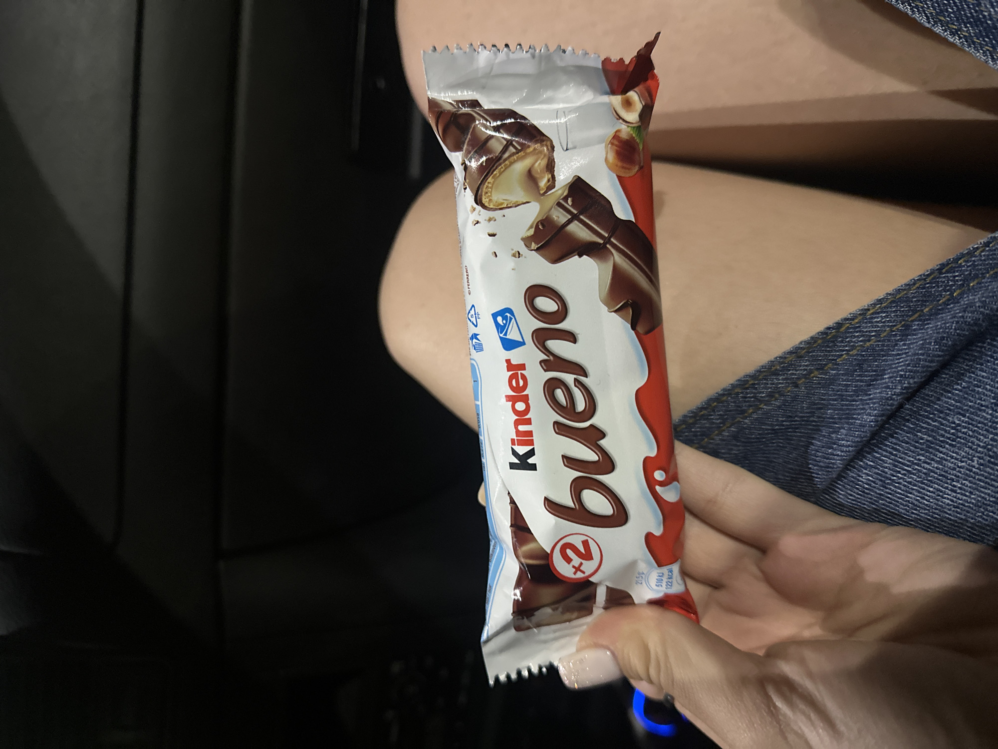 A person's hand, with a manicured fingernail, holds a Kinder Bueno chocolate bar. The chocolate bar is partially unwrapped, revealing the creamy filling and hazelnut pieces. The person is wearing denim clothing. The background is dark and appears to be the interior of a vehicle.