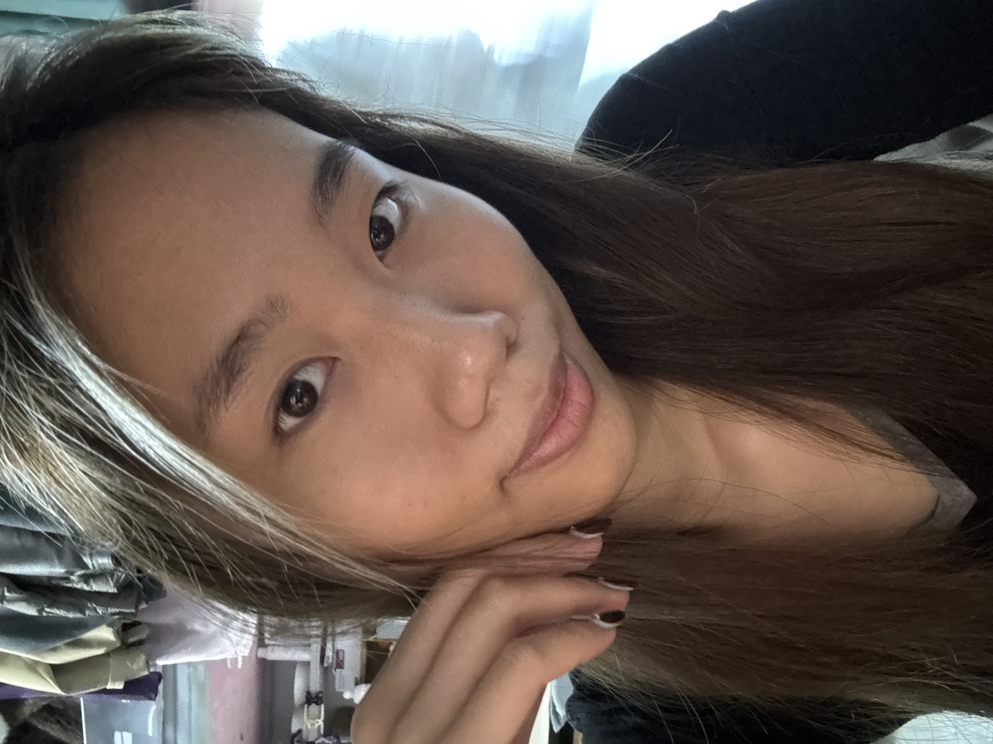 A selfie photograph shows an Asian woman indoors. She has dark brown hair with blonde streaks and brown eyes. She is wearing a dark-colored top. Her right hand is resting on her chin. The background is blurred, but appears to be a cluttered interior with various objects. The image does not provide any information to determine the location to be Nan'an District, China.