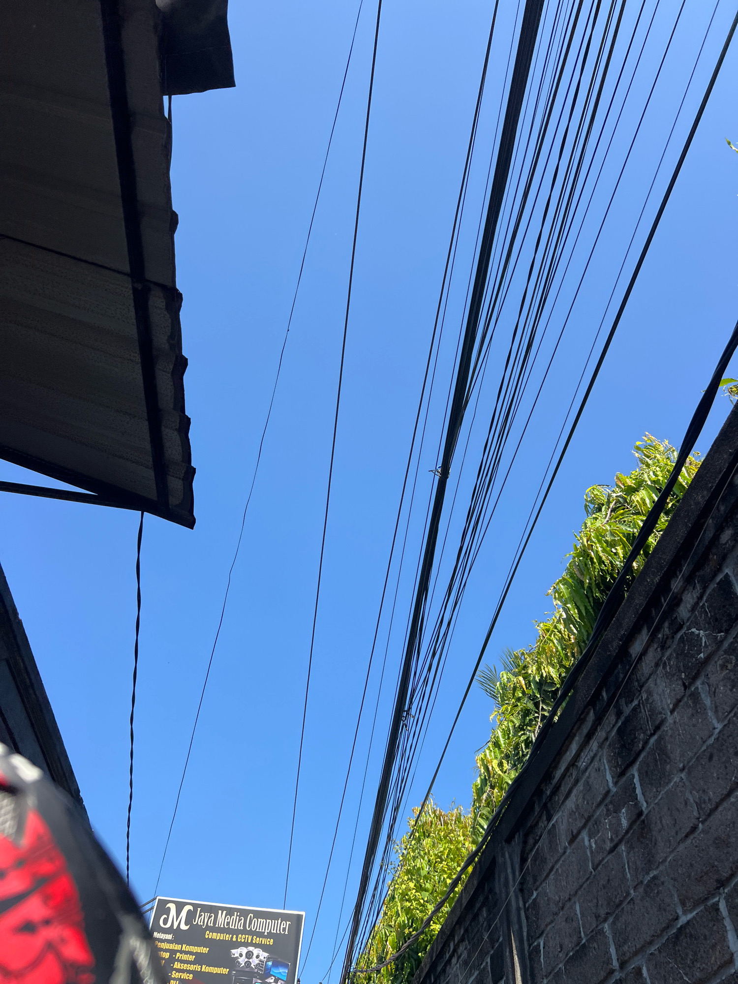 The image shows a view looking upwards towards a clear blue sky, with a dense network of black electrical wires dominating the upper portion of the frame. On the left, a dark, corrugated metal overhang is visible, casting a shadow. To the right, a rough, grey brick wall runs along the bottom edge of the frame, topped with lush green foliage. A sign for "Jaya Media Computer" is partially visible near the bottom center, advertising computer and CCTV services. The scene suggests an urban environment, likely a street in Kuta, Indonesia, given the typical overhead wiring and architectural elements. There are no people or discernible actions depicted in the image.