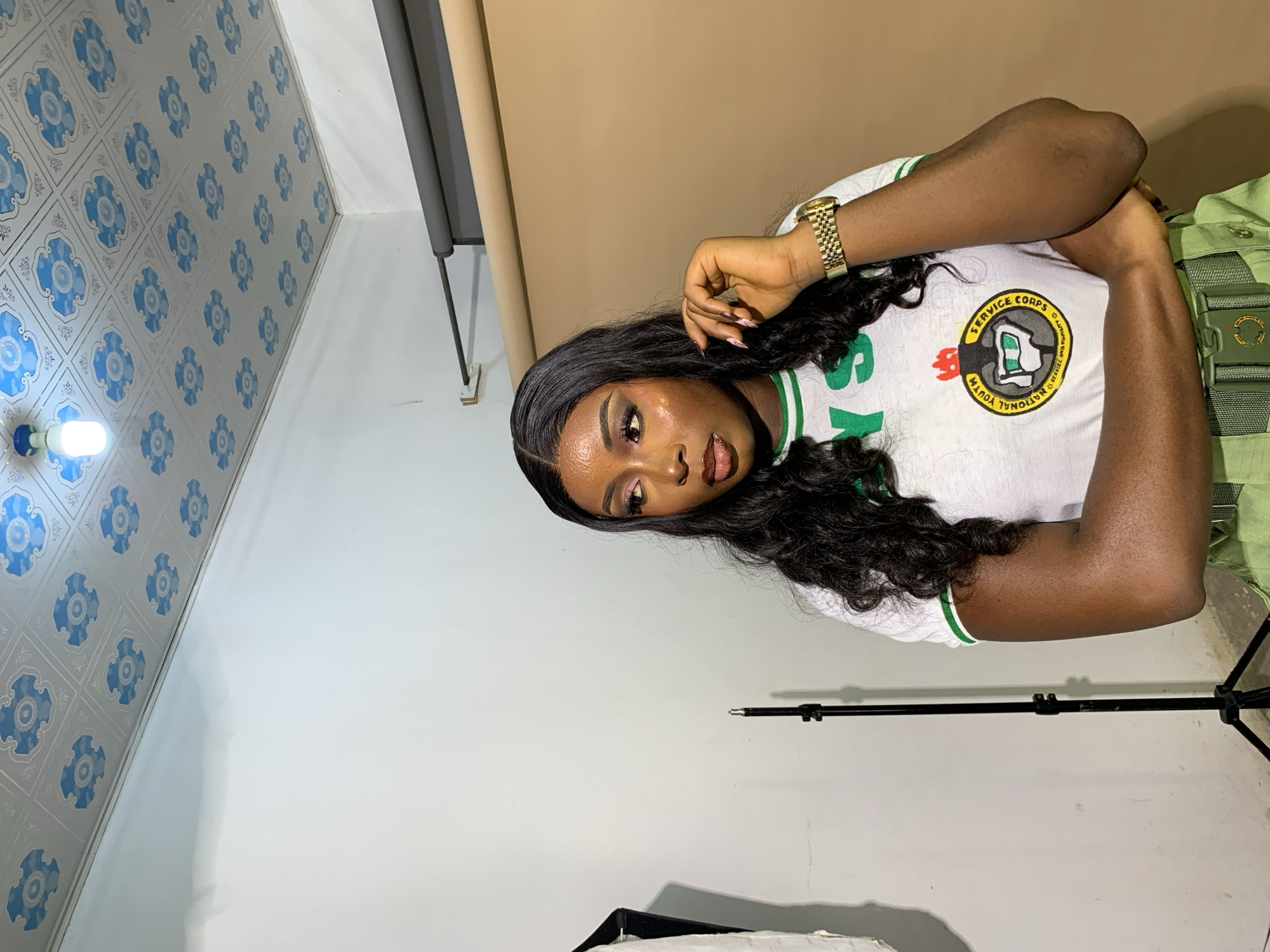 In a studio setting, a young Black woman with dark, wavy hair and dramatic makeup stands with her arms crossed. She is wearing a white t-shirt with green trim and "NYS" printed in green letters, along with a circular emblem featuring the Nigerian National Youth Service Corps logo. She also wears a green belt over green pants, suggesting a uniform. Her right hand is raised to her face, and she looks off to the side. In the background, a white wall transitions to a tan backdrop, with studio lighting equipment and a patterned ceiling visible. The scene is devoid of any specific indicators of Gura Top, Nigeria.