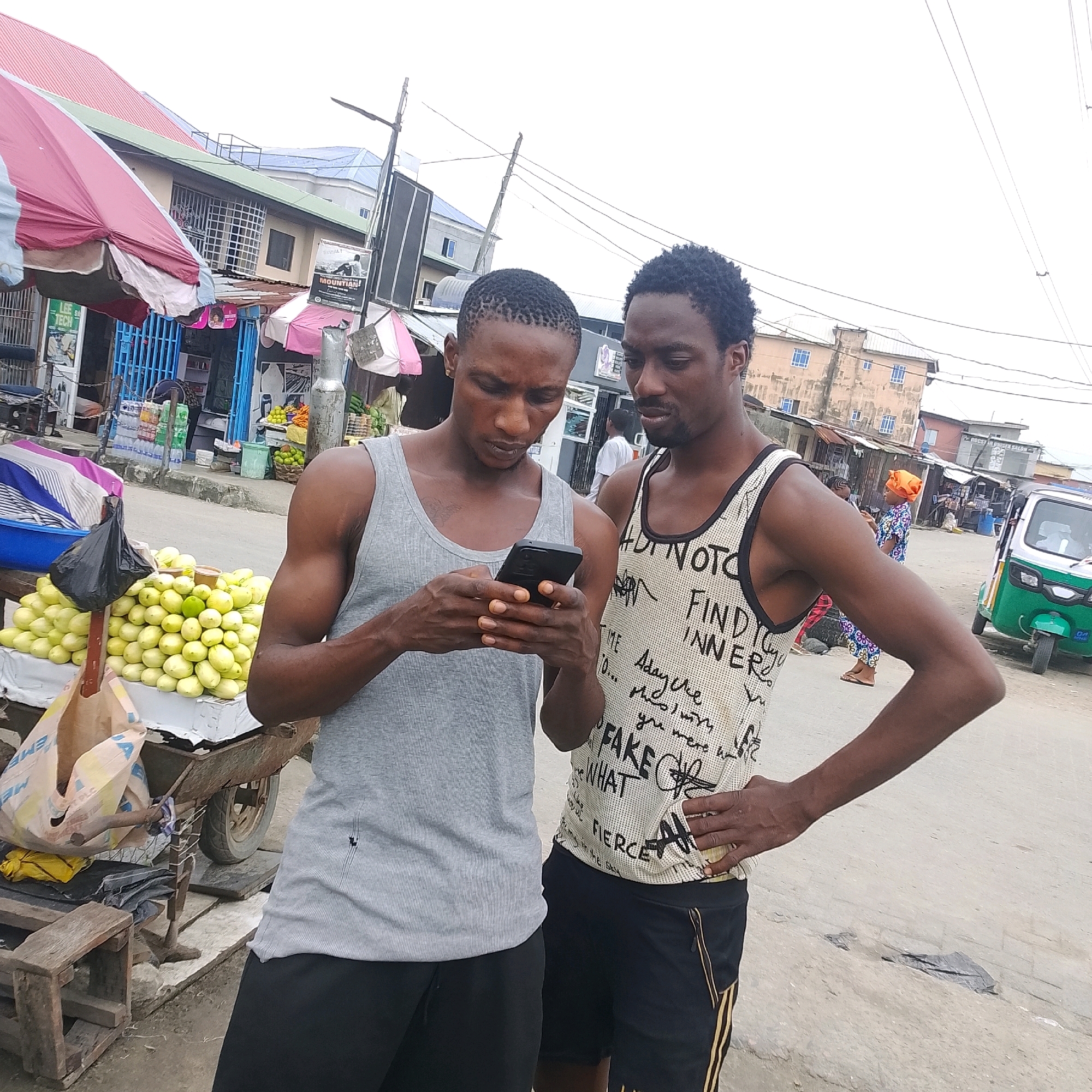 In Ado, Nigeria, two young men stand on a dusty street in front of market stalls. The man on the left, wearing a grey tank top and black shorts, is holding a smartphone and looking down at it. The man on the right, sporting a patterned tank top and black shorts, stands close to him, also looking at the phone. Behind them, a vibrant street scene unfolds with various market stalls laden with produce, including piles of green fruits. Colorful umbrellas provide shade, and buildings with barred windows line the street. A small, green and white motorized tricycle is parked nearby, and in the background, other market-goers and shoppers are visible, creating a bustling atmosphere. The image captures a candid moment of everyday life in this Nigerian city.