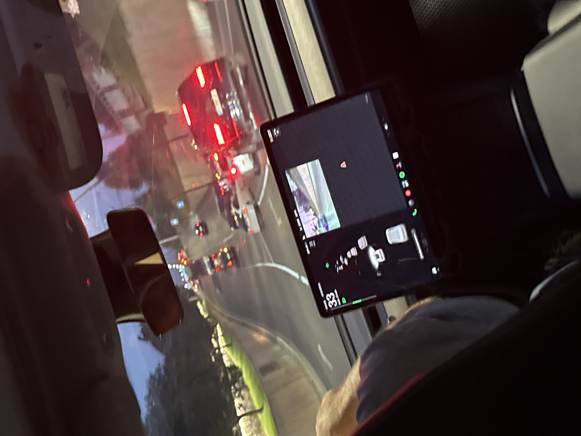 The media file depicts a view from inside a vehicle, likely a car, looking through the windshield. The scene outside appears to be a city street with significant traffic. Red taillights of vehicles are prominent, suggesting it is either evening or there is low visibility. The road is curved, and there are lights from streetlamps and buildings visible along the sides. A digital tablet is mounted on the dashboard, displaying what appears to be a camera feed, possibly from the vehicle's front camera, along with some interface elements and a time display of "1:33". The reflection on the windshield indicates a somewhat overcast or evening sky. A person's arm and shoulder are visible in the foreground, suggesting they are a passenger or the driver. The location is identified as Tbilisi, Georgia. The overall atmosphere is that of being in transit, possibly stuck in traffic.