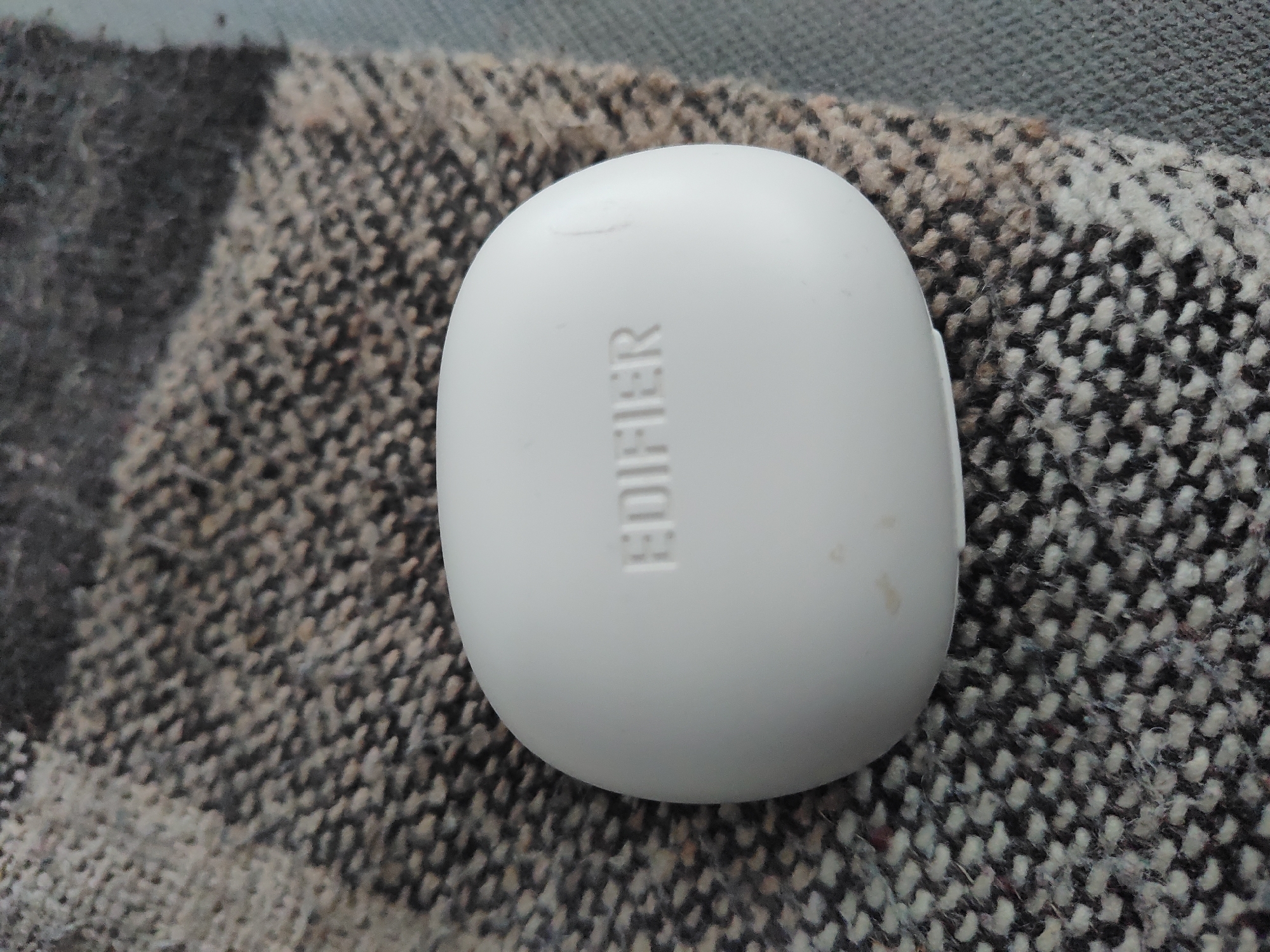A white, oval-shaped Edifier earbud case sits on a patterned, woven fabric in an indoor setting. The fabric has a repeating pattern of dark and light shades, creating a textured appearance. The location is unknown and is not identifiable as Kaspichan, Bulgaria. No people, interactions, or actions are visible.
