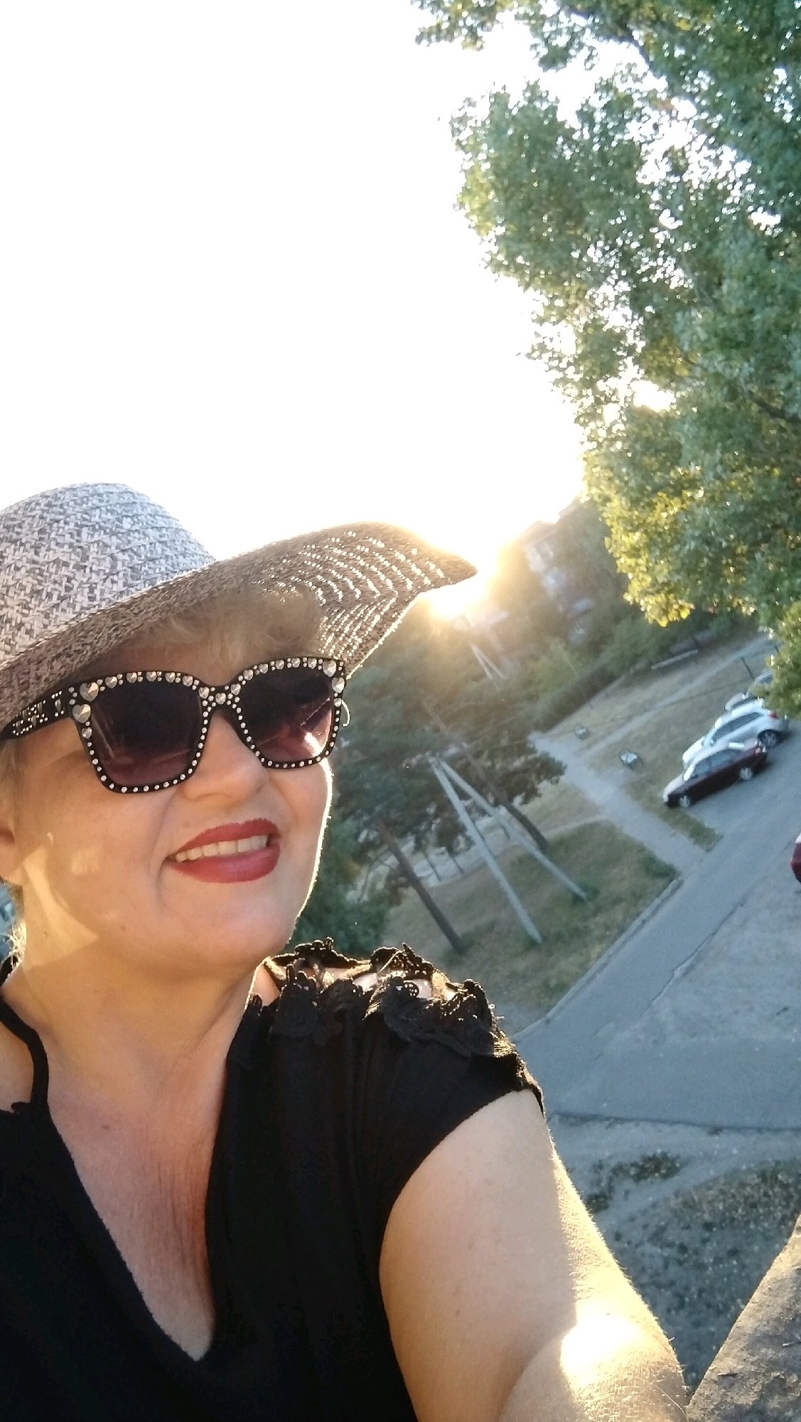 A woman wearing a patterned hat and sunglasses smiles at the camera. She is wearing a black top with lace accents on the shoulders. In the background, there are trees and buildings, suggesting an outdoor urban setting. The sun is setting, casting a warm glow on the scene. There are two cars parked on a road in the distance. The location is identified as Horishni Plavni, Ukraine.