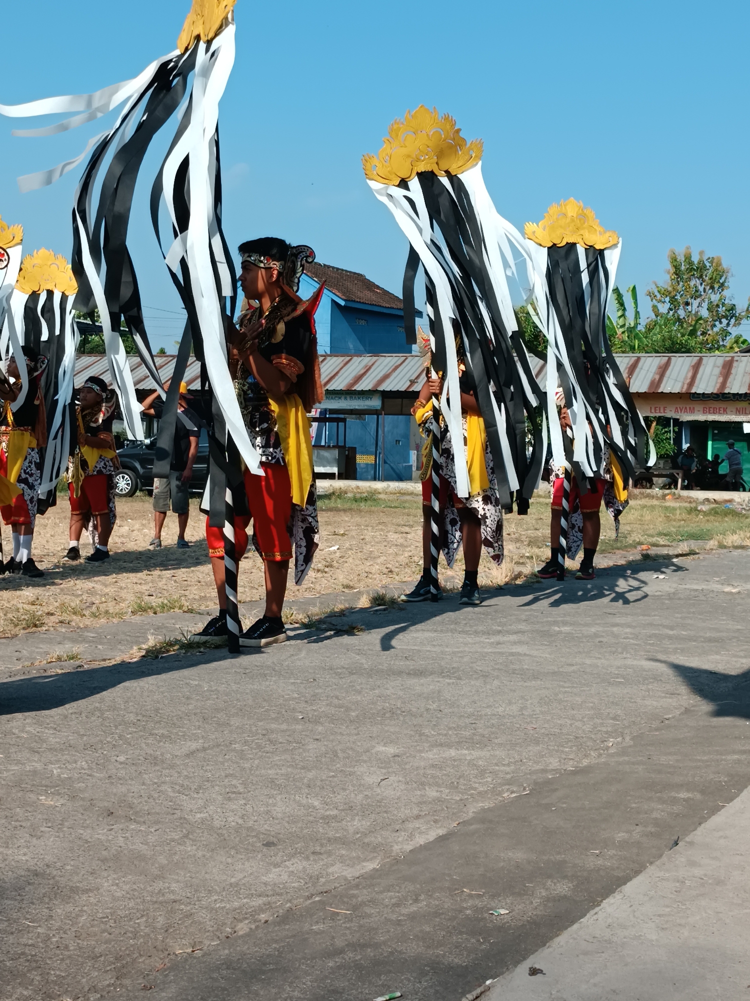 In Imogiri, Indonesia, on a bright, clear day with a blue sky, several individuals are participating in a procession. They are dressed in traditional attire, featuring red shorts, yellow tops, and elaborately decorated headwear and costumes adorned with black and white ribbons that cascade downwards. The procession appears to be moving along a paved walkway adjacent to a dry, grassy area. In the background, a blue building with a corrugated metal roof stands, and further beyond, another building with a similar roof displays a sign that reads "LELE AYAM BEBEK - NILU." Several trees are visible in the distance. One of the participants in the foreground is holding a staff-like object with black and white ribbons. The shadows cast by the individuals and their regalia suggest the sun is high in the sky.