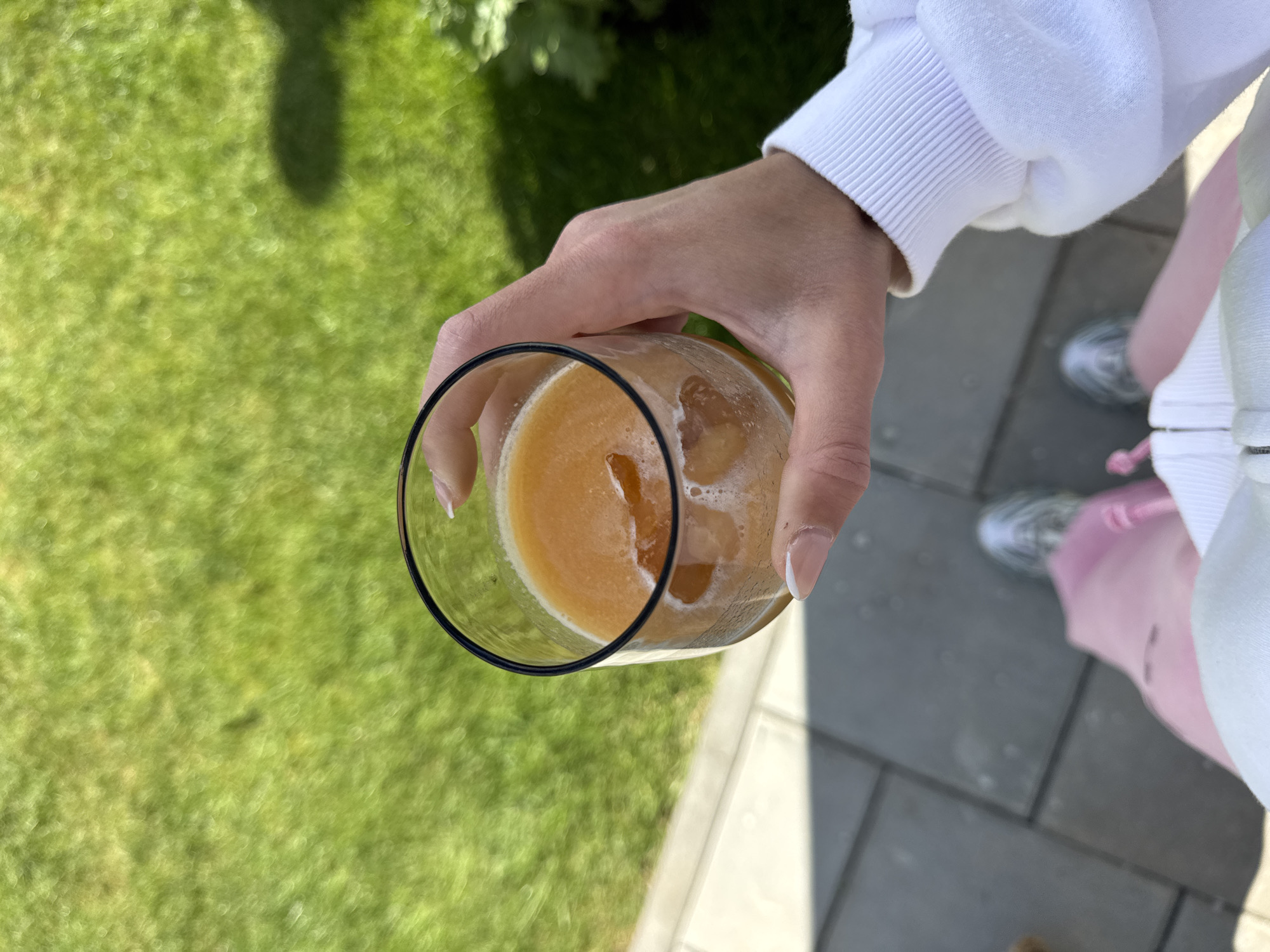 A first-person, high-angle shot depicts a hand holding a glass of orange liquid with ice. The hand belongs to a person wearing a white long-sleeved top. The background consists of green grass, gray paving stones, and a glimpse of another person's leg, pink fabric, and gray and white sneakers. There is no verifiable information to confirm this photo was taken in Blagoevgrad, Bulgaria.