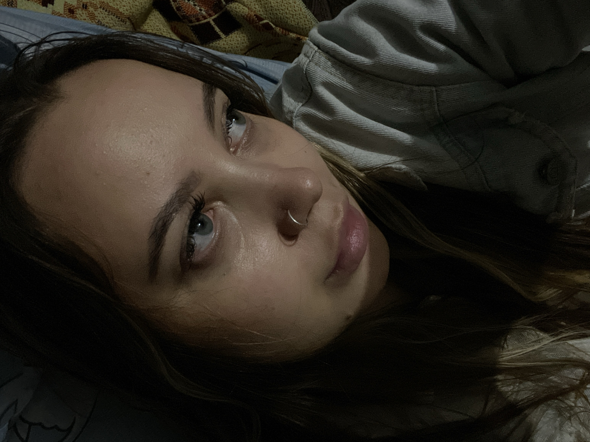A young woman with light eyes and a nose ring is lying down. She is wearing a light-colored, buttoned shirt. The background consists of bedding. The location is not evident from the image. The city of Novopokrovskaya, Russia, cannot be determined.
