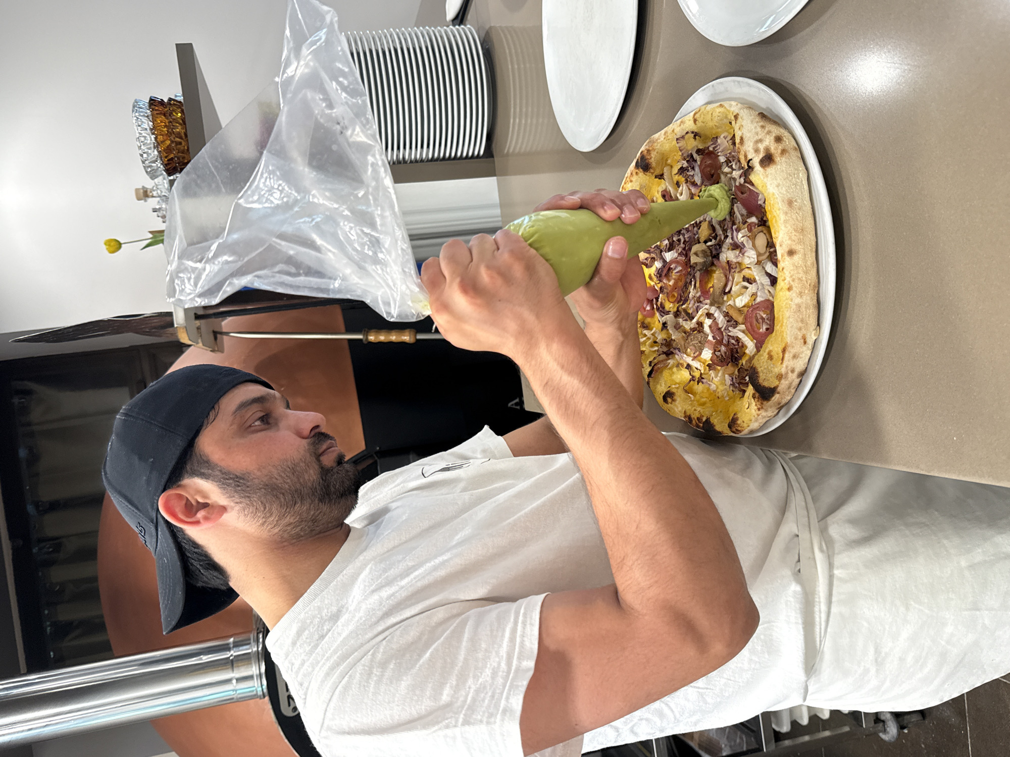 A person wearing a white t-shirt and apron is piping a green sauce onto a pizza. The person is wearing a black baseball cap. The pizza is on a white plate. In the background, there is a pizza oven, a shelf with dishes and a yellow flower, and a stack of white plates. The scene appears to be a restaurant or kitchen in Trento, Italy.