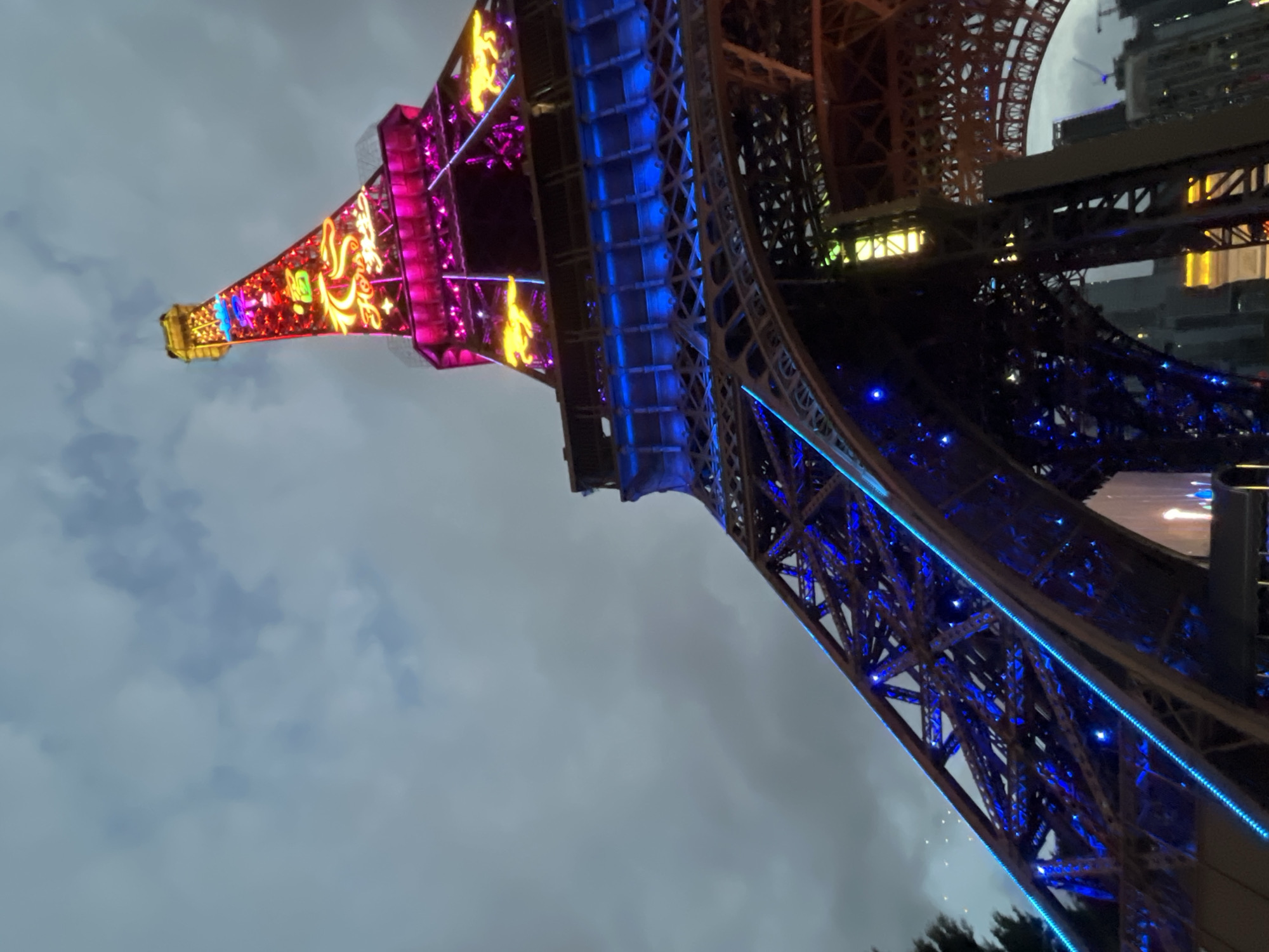 A low-angle shot depicts a brightly lit replica of the Eiffel Tower against a cloudy sky, likely located in the city of Nanshan District, China. The tower is illuminated with a colorful array of lights, including pink, red, yellow, and blue hues. The base of the tower is primarily blue, transitioning to pink and red with ornate designs as it ascends. Adjacent buildings are visible.